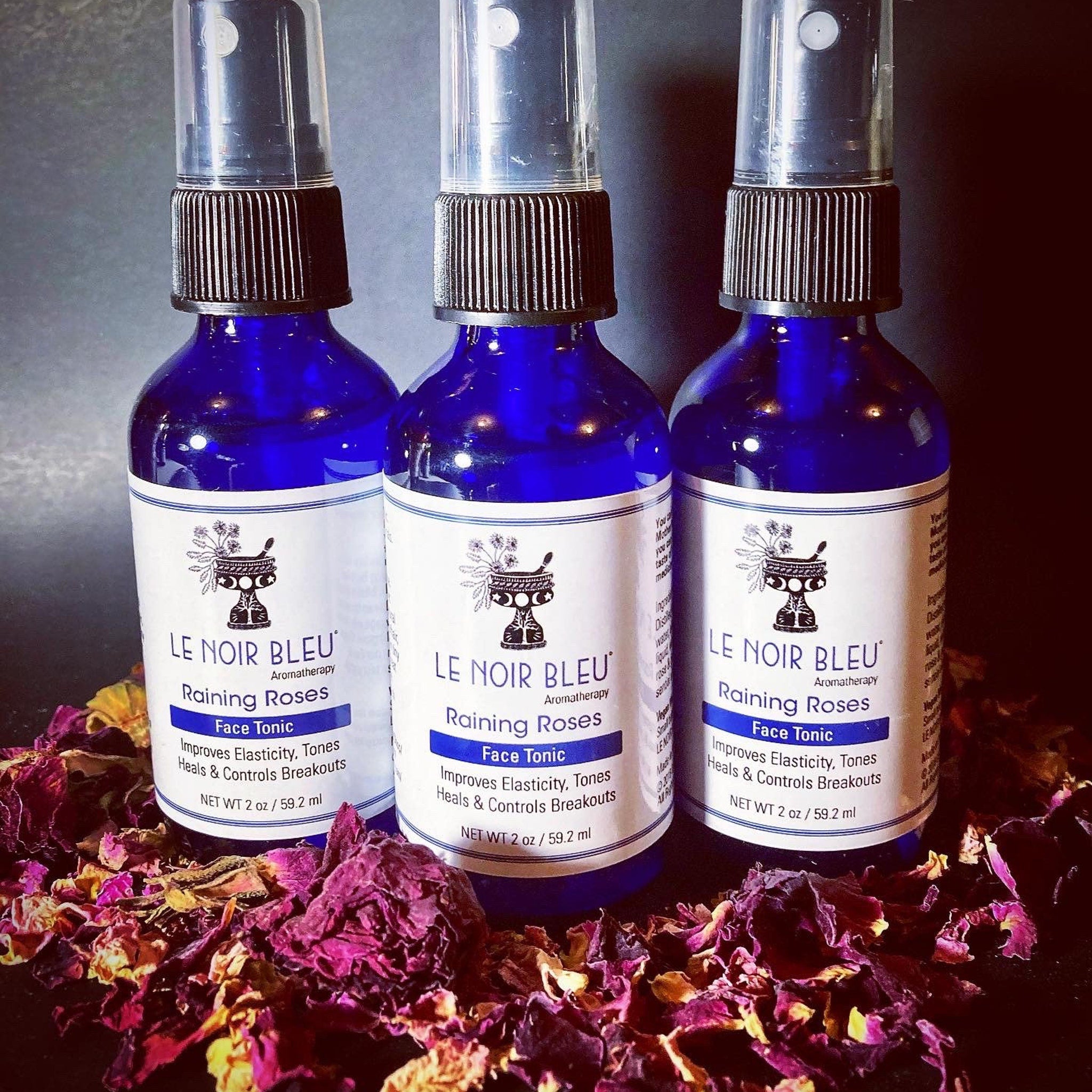 Face tonic bottles with blue glass, black caps, and white labels with "LE NOIR BLEU" text, surrounded by rose petals.