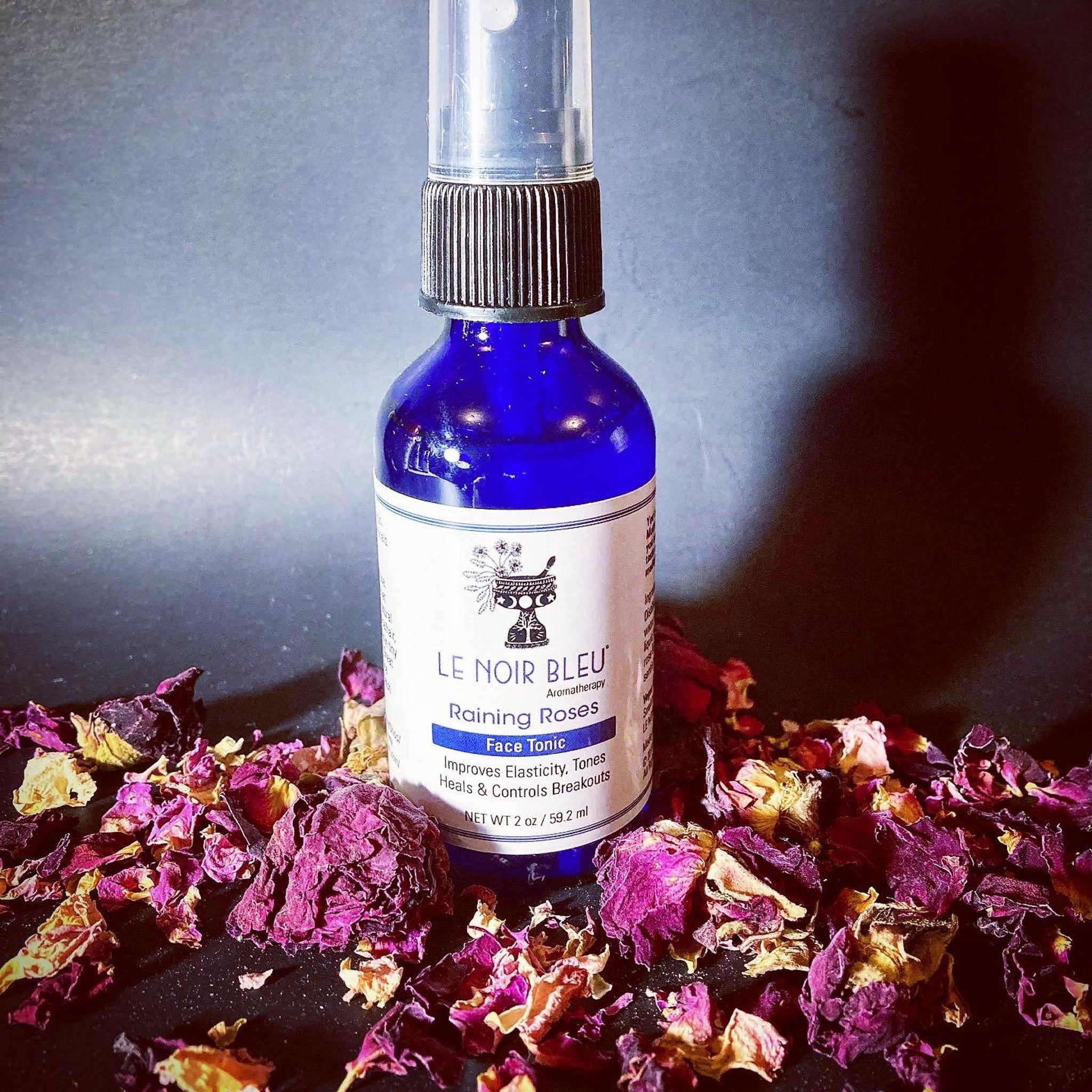 Face tonic bottle with blue glass, black cap, and "LE NOIR BLEU" label, surrounded by rose petals