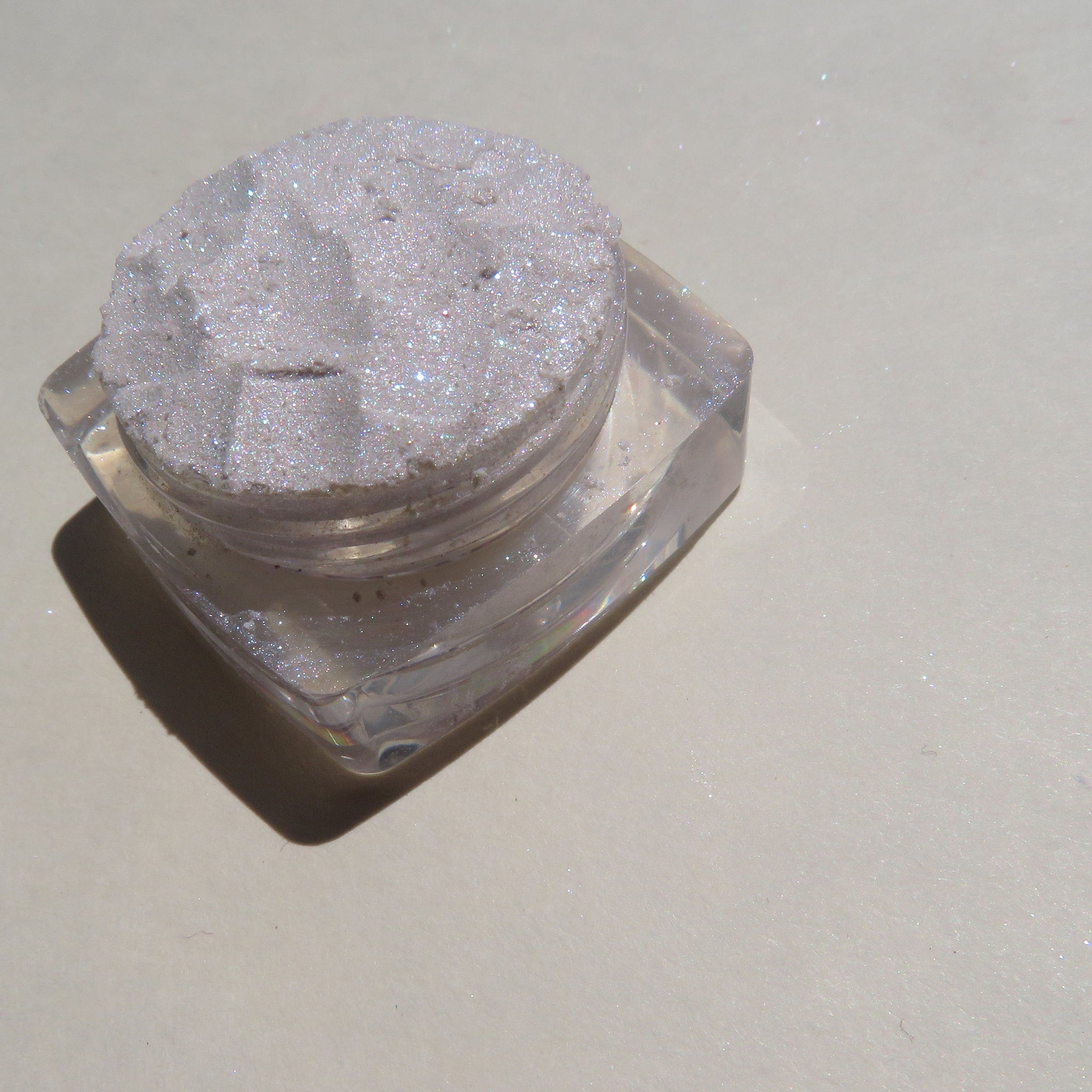White loose pigment eyeshadow with violet shimmer in a clear jar, showcasing sparkly texture on a pink surface.