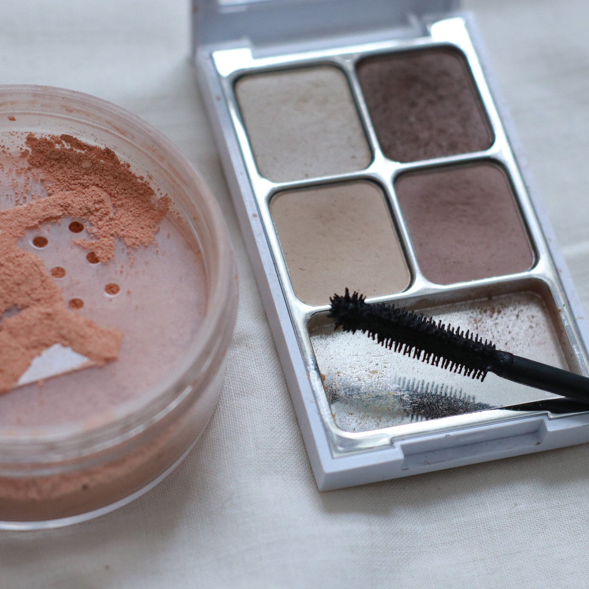 Eyeshadow palette with brown, beige, and tan shades, black mascara brush, and pink loose powder in a clear container