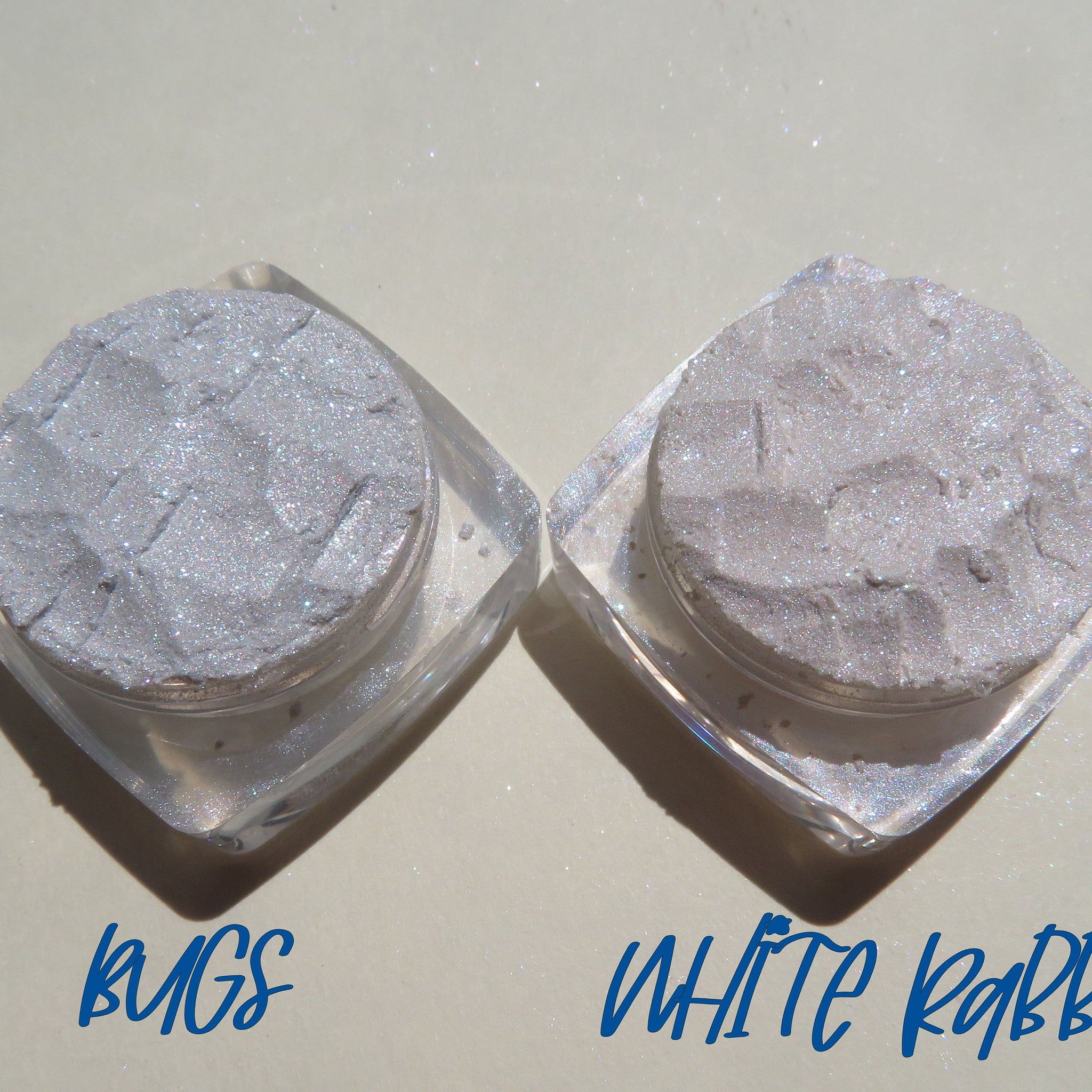 Two open jars of loose eye shadow pigments in white and taupe with shimmer particles, displayed on a reflective surface.