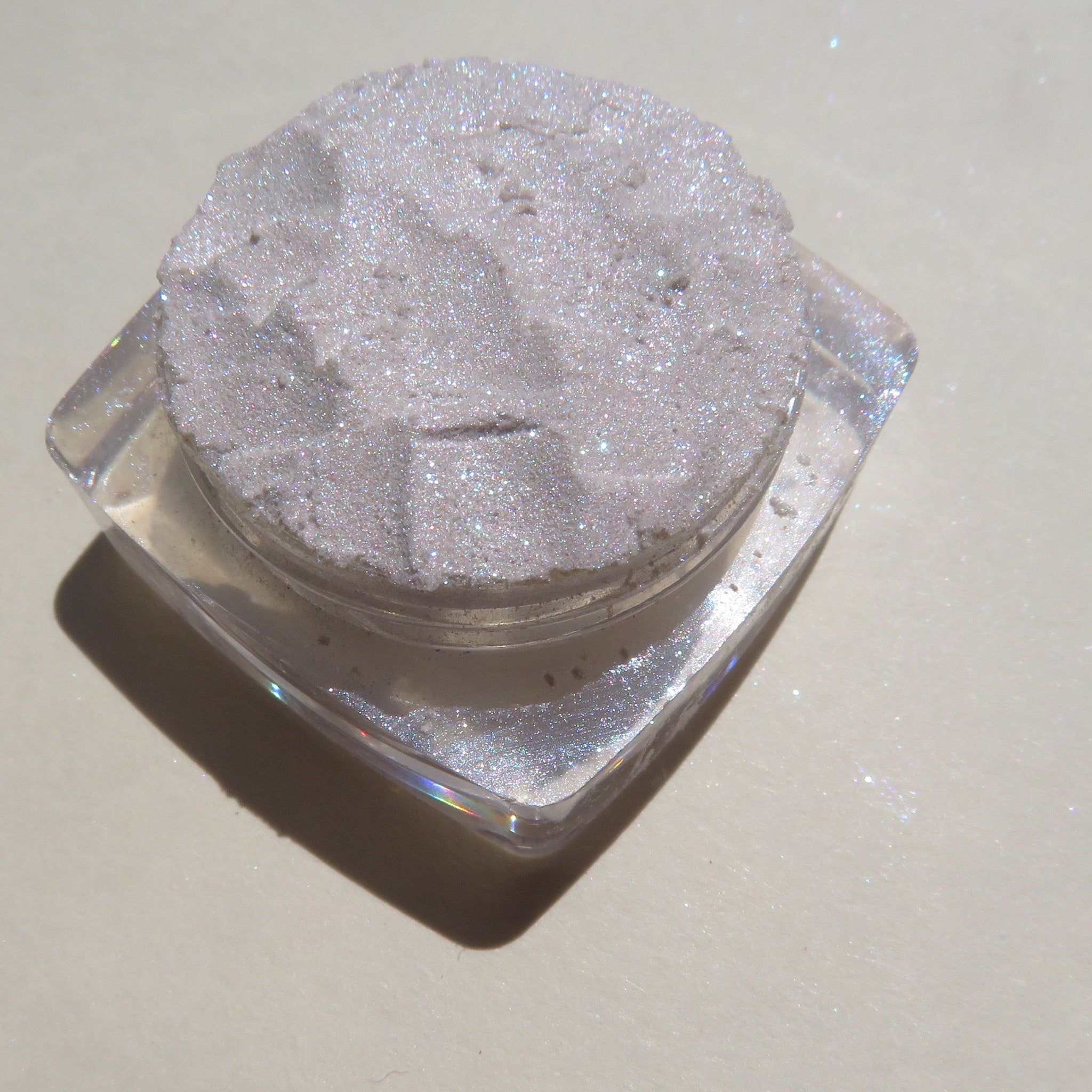 Side view of loose white eyeshadow with violet shimmer in a clear jar, showcasing sparkly glitter particles.