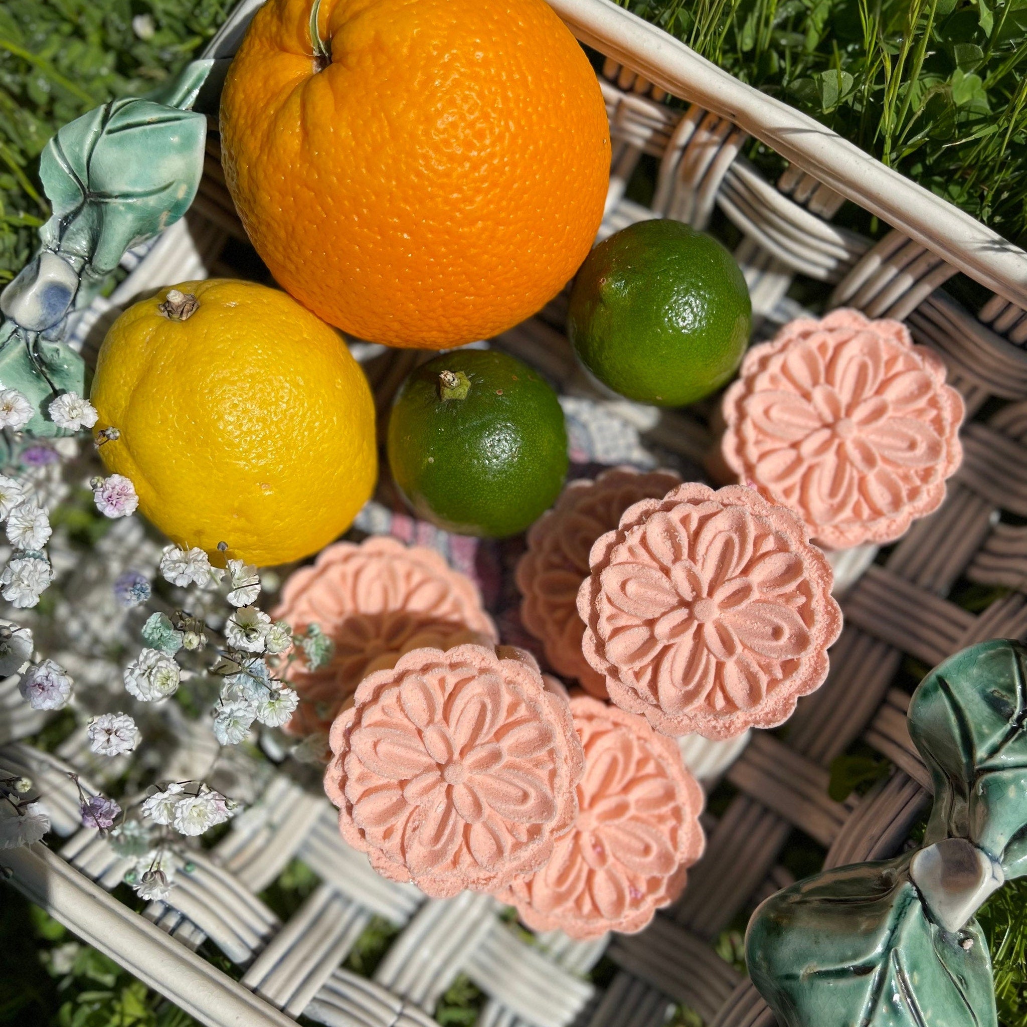Pink floral shaped soaps with orange, lemon, and limes in a woven basket on grass