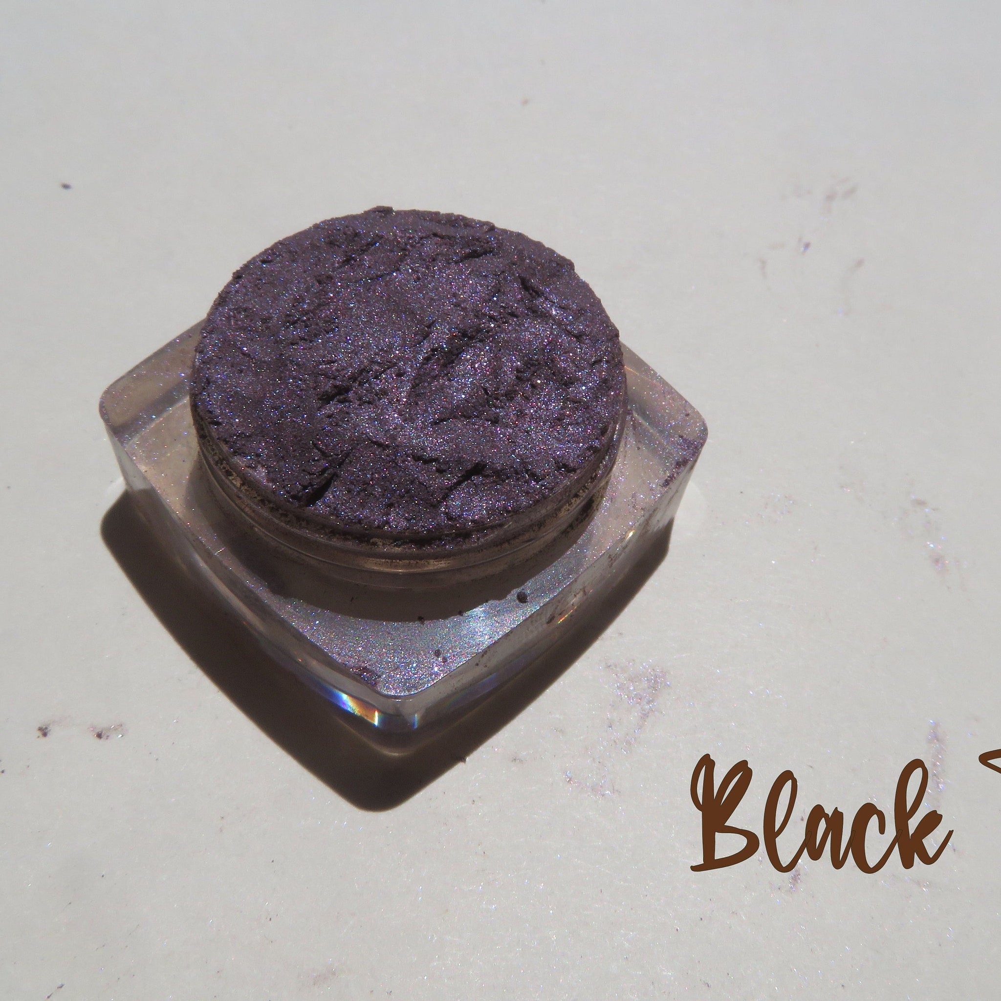 Side view of loose mineral eyeshadow jar showing dark black purple shimmer powder spilling slightly