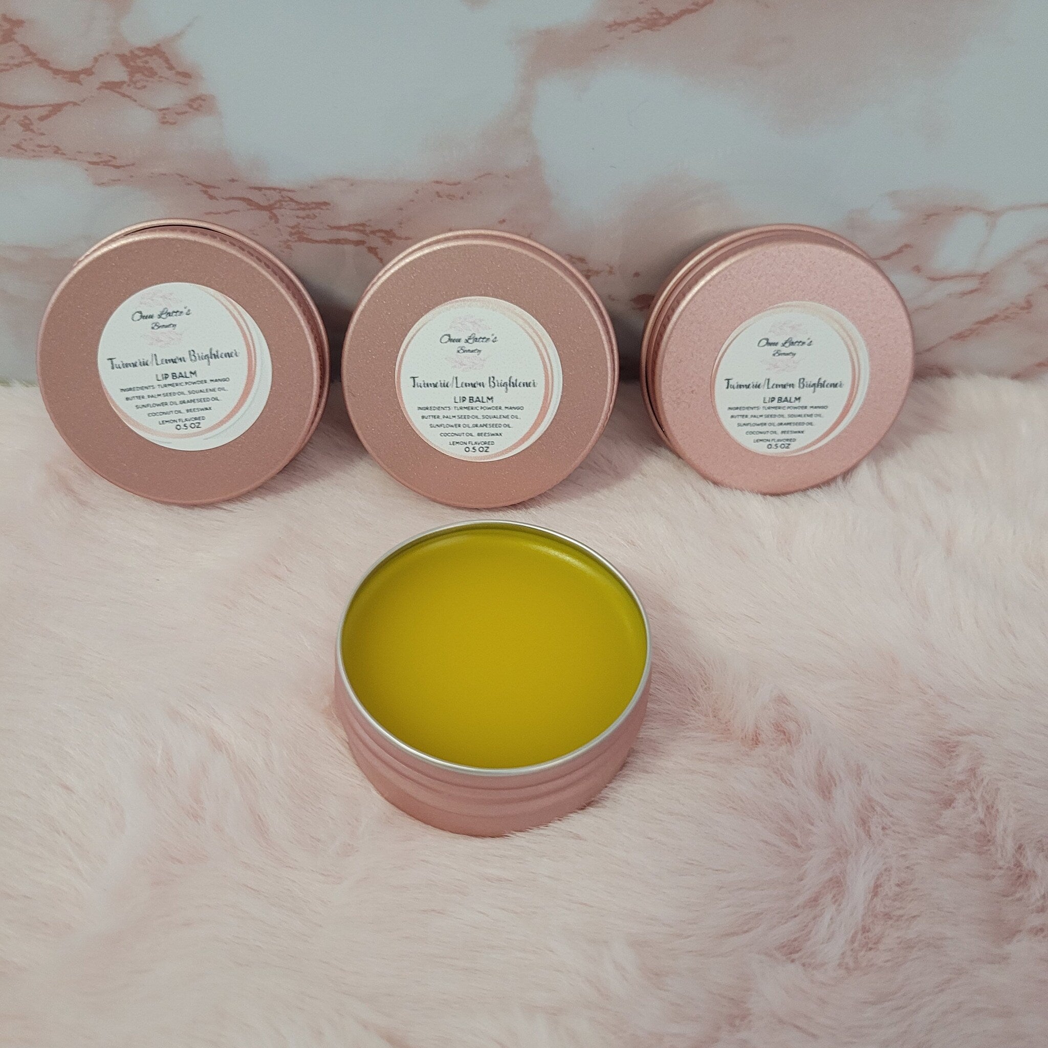 Lip balm in pink metal tins with yellow balm, on pink fluffy surface, by Ona Latte's Beauty