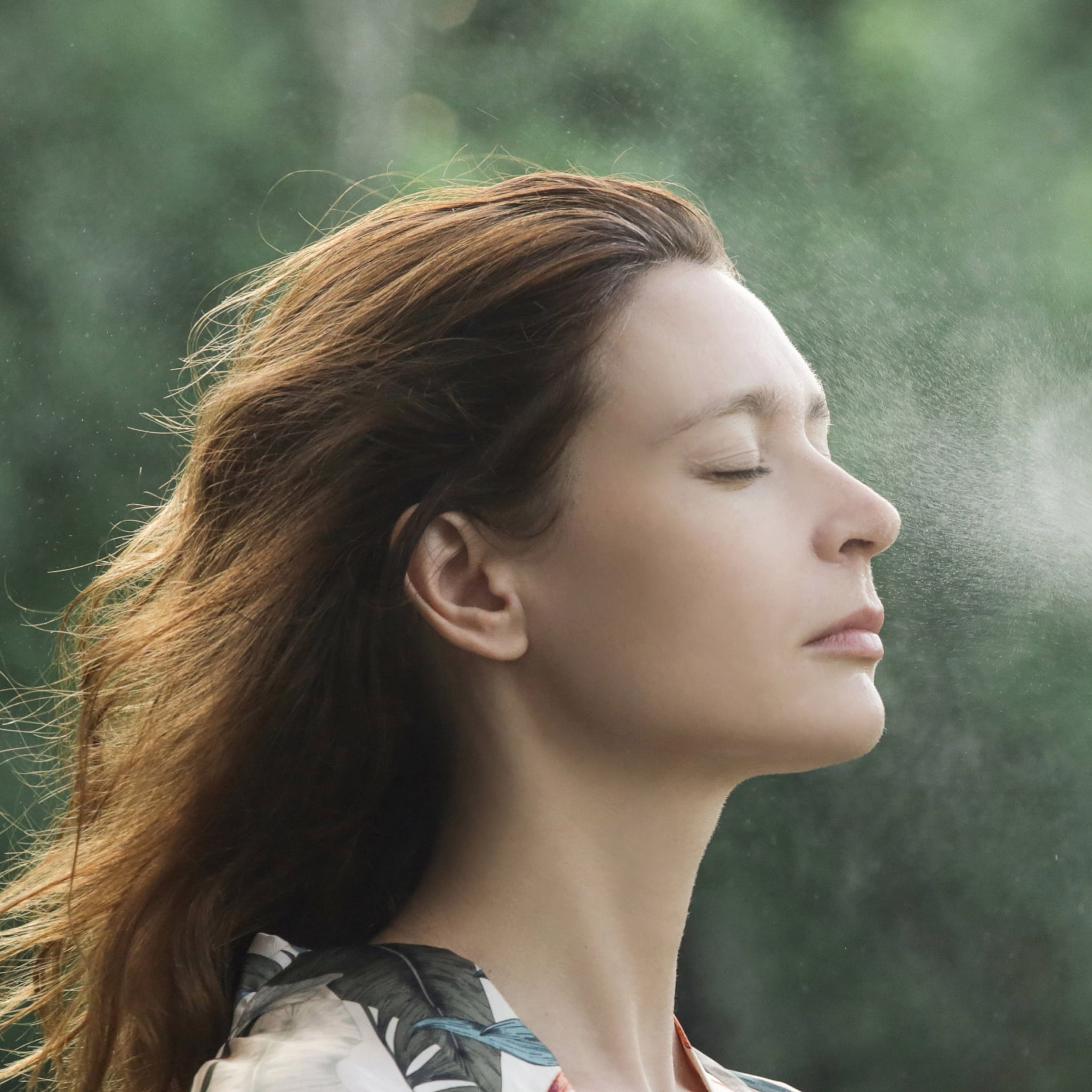 Woman with auburn hair and closed eyes, misting face with water spray, blurred green background