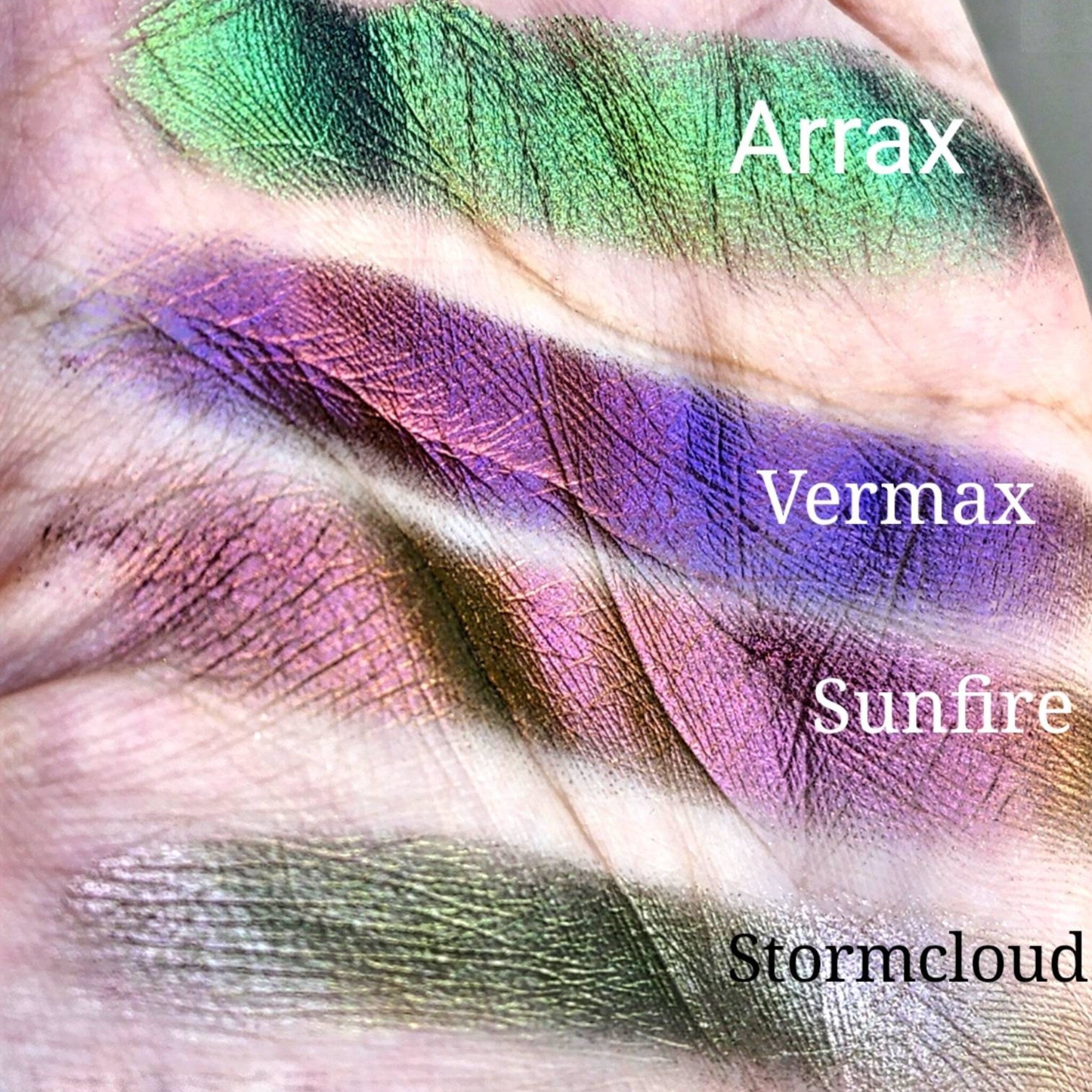 Swatches of green, purple, and pink iridescent eyeshadow on skin, with text labels Arrax, Vermax, Sunfire, and Stormcloud