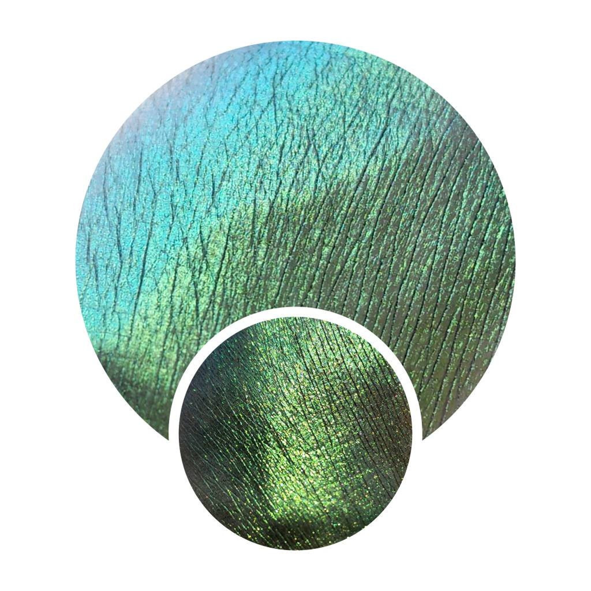 Eye shadow swatch with green and blue color shift, textured finish, and sparkle effect