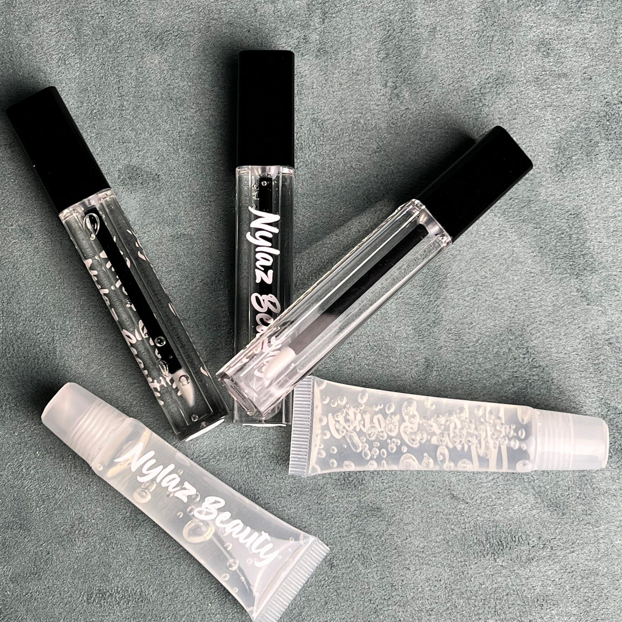 Lip gloss tubes, clear plastic, black caps, with Nylaz Beauty text, on gray surface