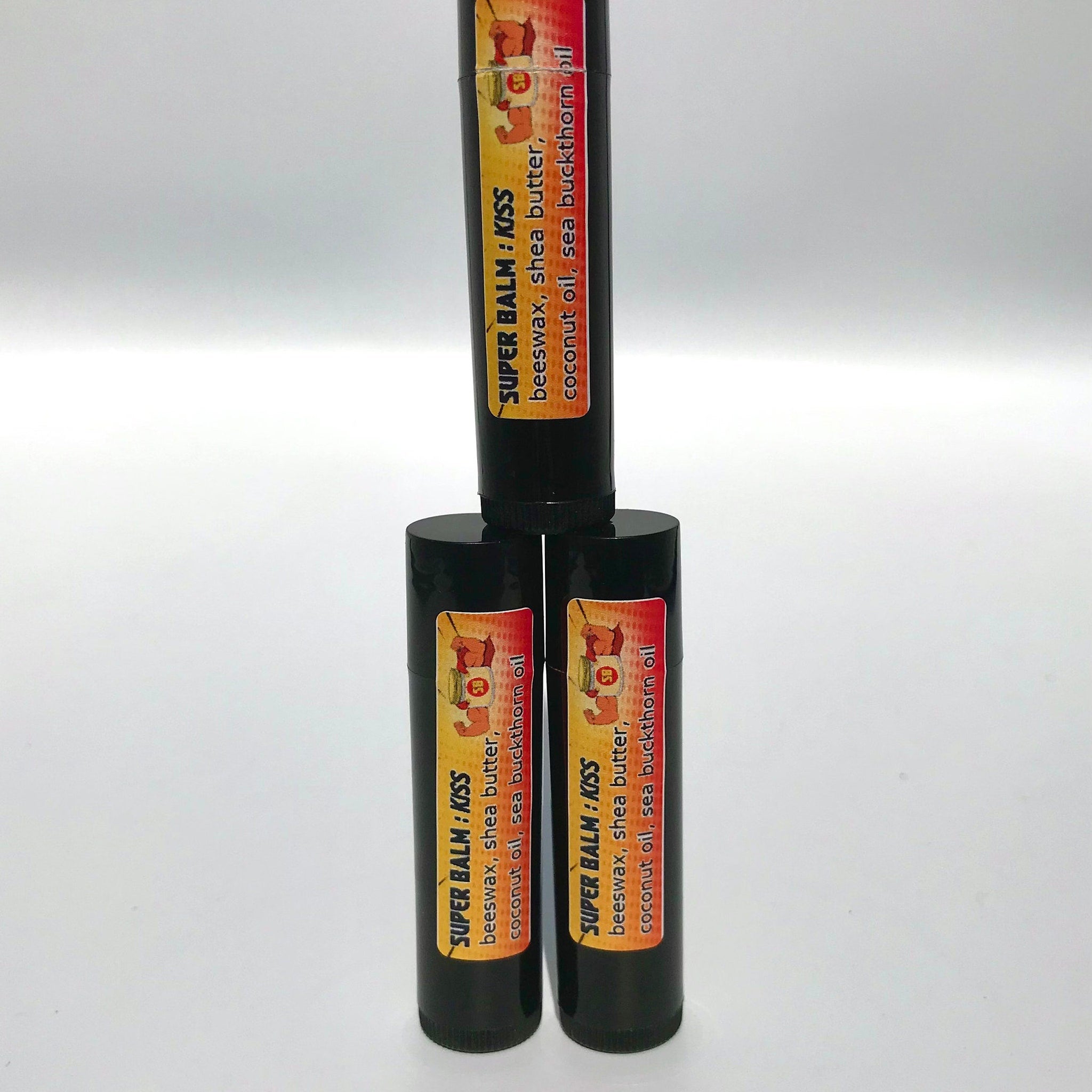 Lip balm tubes with black casing and orange/yellow label, stacked together, "SUPER BALM: KISS" text visible