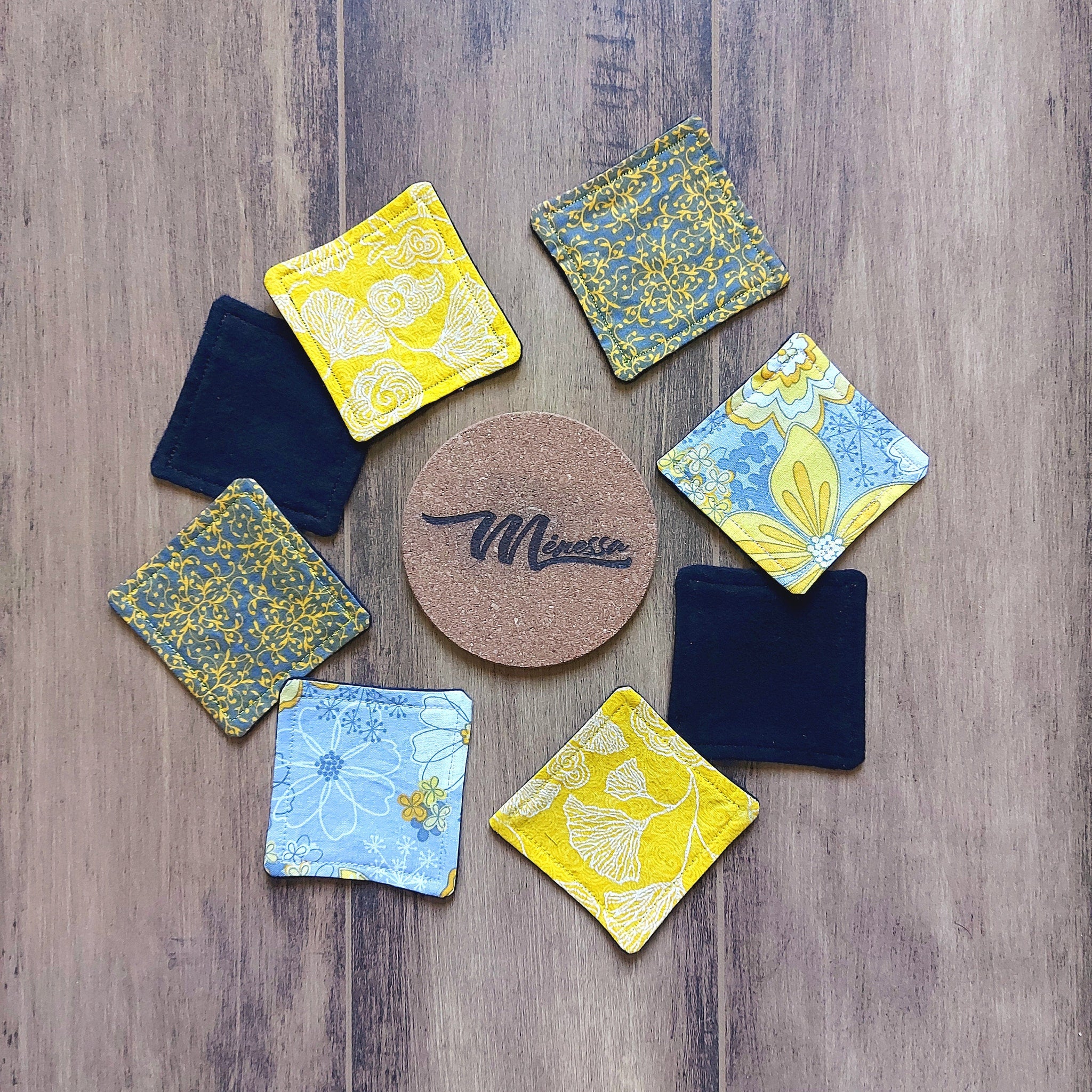 Coasters with floral patterns, in yellow, blue, and black, with cork center and "Mènessa" brand logo