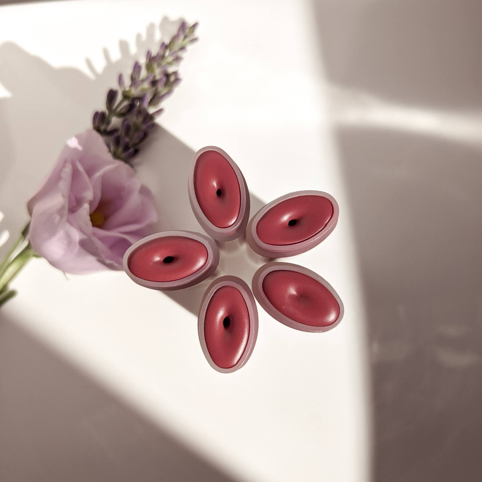 Lip balm, pink oval-shaped with dark center, arranged with flower and lavender sprig on white surface