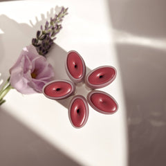 Lip balm, pink oval-shaped with dark center, arranged with flower and lavender sprig on white surface