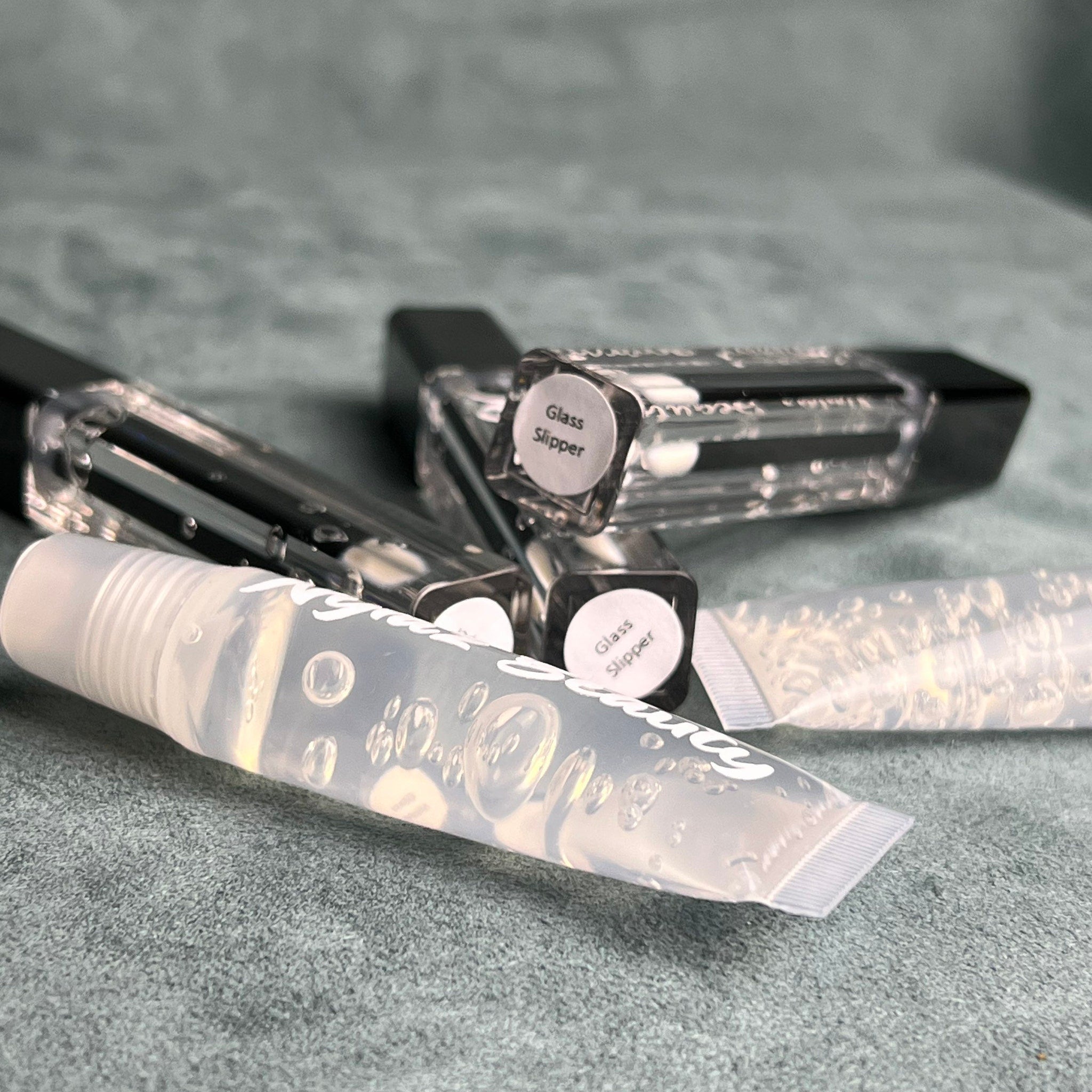 Lip gloss tubes, clear with black caps, "Glass Slipper" text, on green fabric background