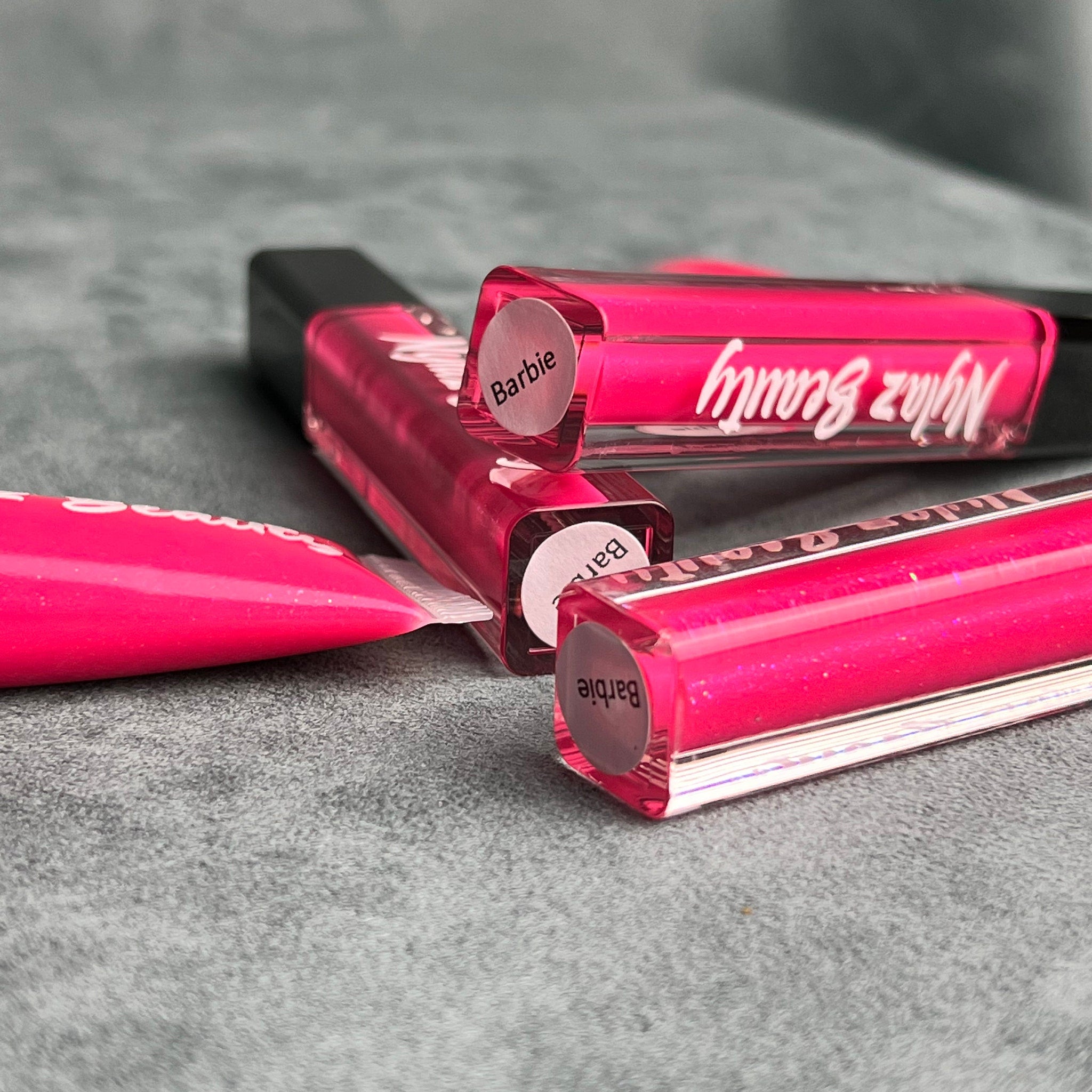 Lip gloss tubes in bright pink with black caps and Nyiaz Beauty branding on a grey surface