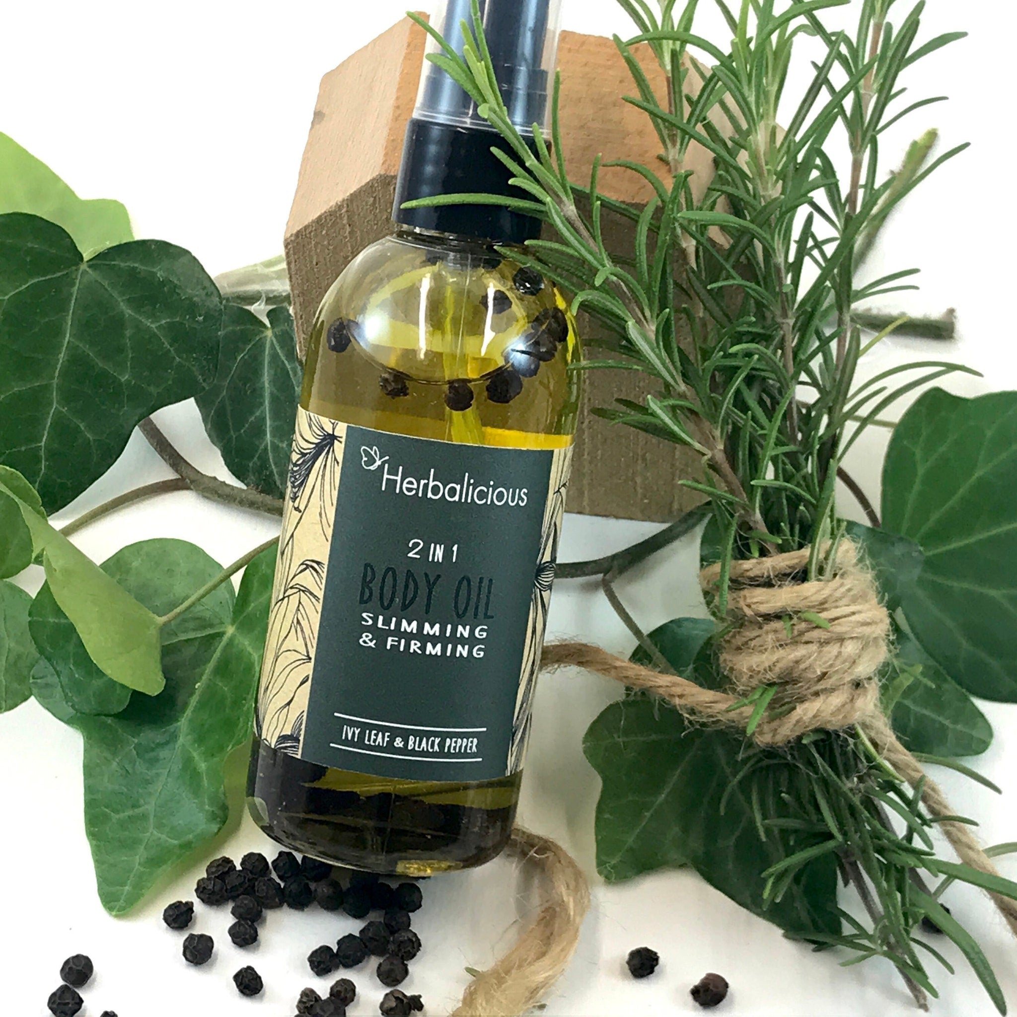 Body oil bottle with Herbalicious label, clear liquid, and black peppercorns, surrounded by green leaves and rosemary sprigs