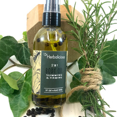 Body oil bottle with green liquid, black peppercorns, and Herbalicious label next to greenery