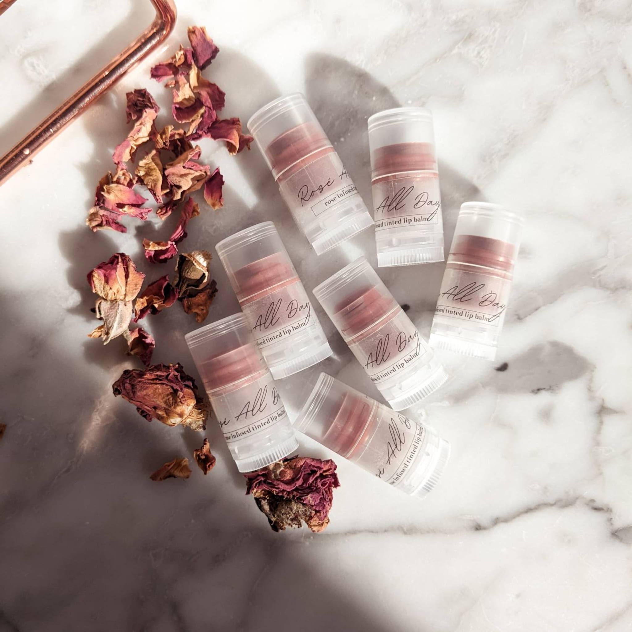 Tinted lip balm tubes with pink balm, clear plastic, and "All Day" text on marble surface