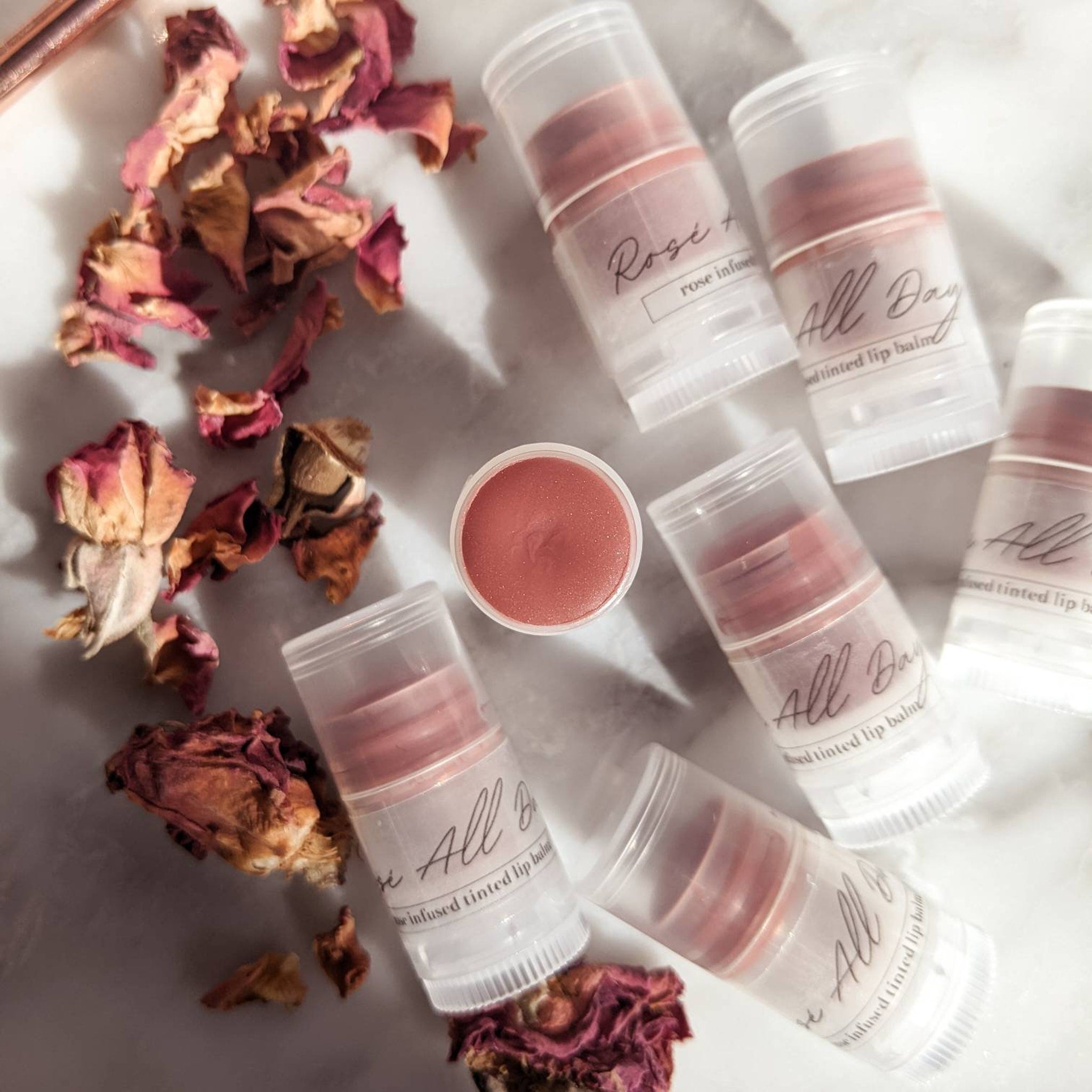 Lip balms in clear tubes with rose-tinted balm, "All Day" text, and dried rose petals