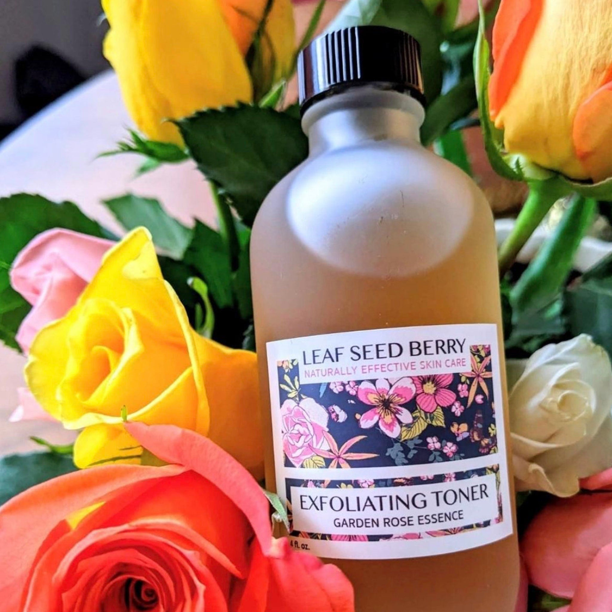 Exfoliating toner bottle, frosted glass, black cap, floral label, surrounded by colorful roses