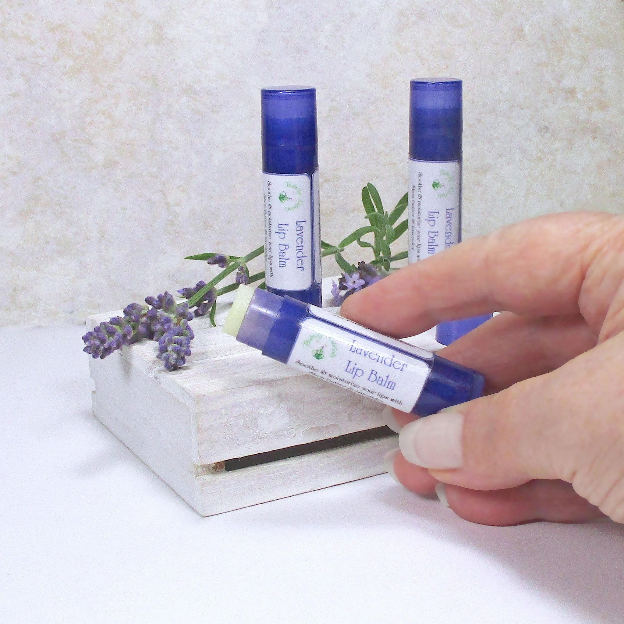 Lavender lip balm in blue tube held by hand, with lavender sprigs on white surface