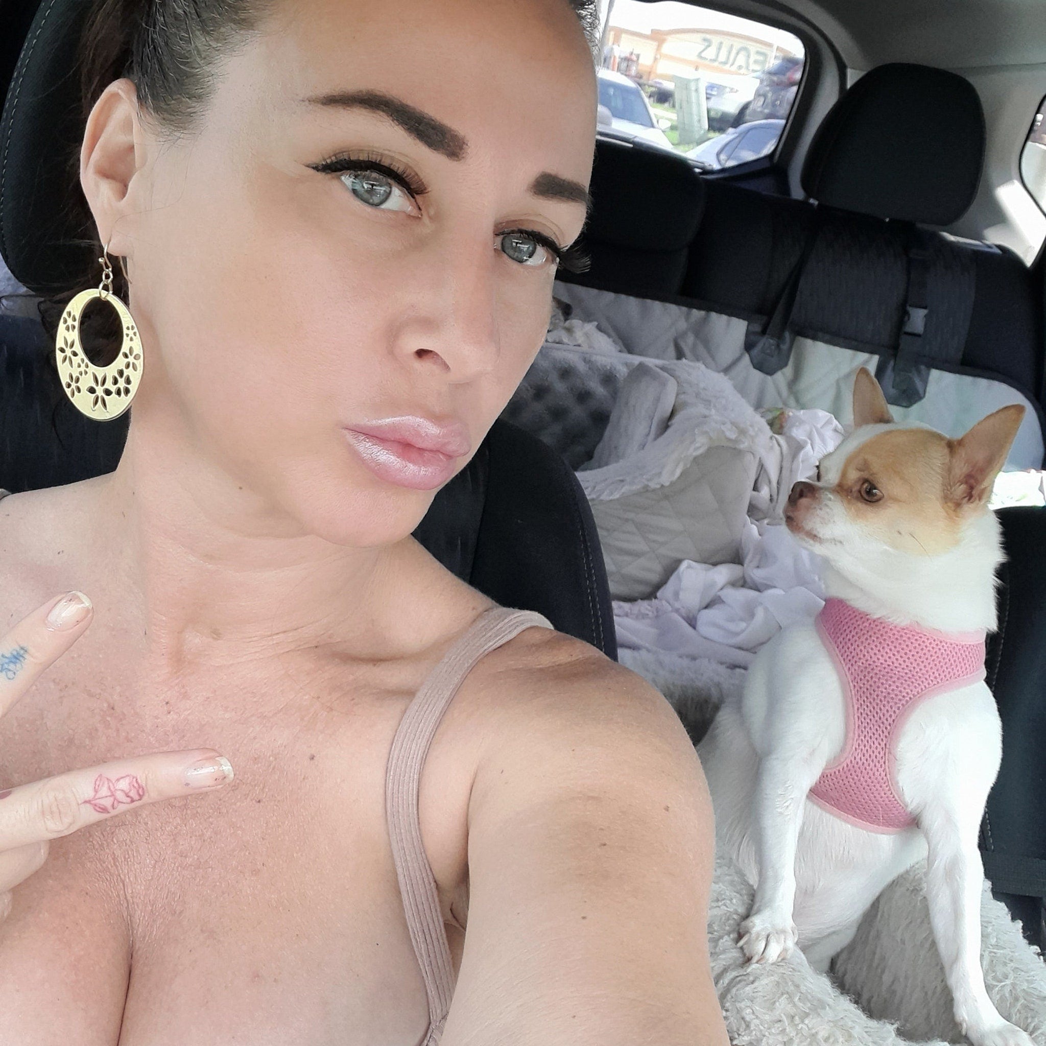 Woman with gold earrings, pink lips, and dog in car with pink harness