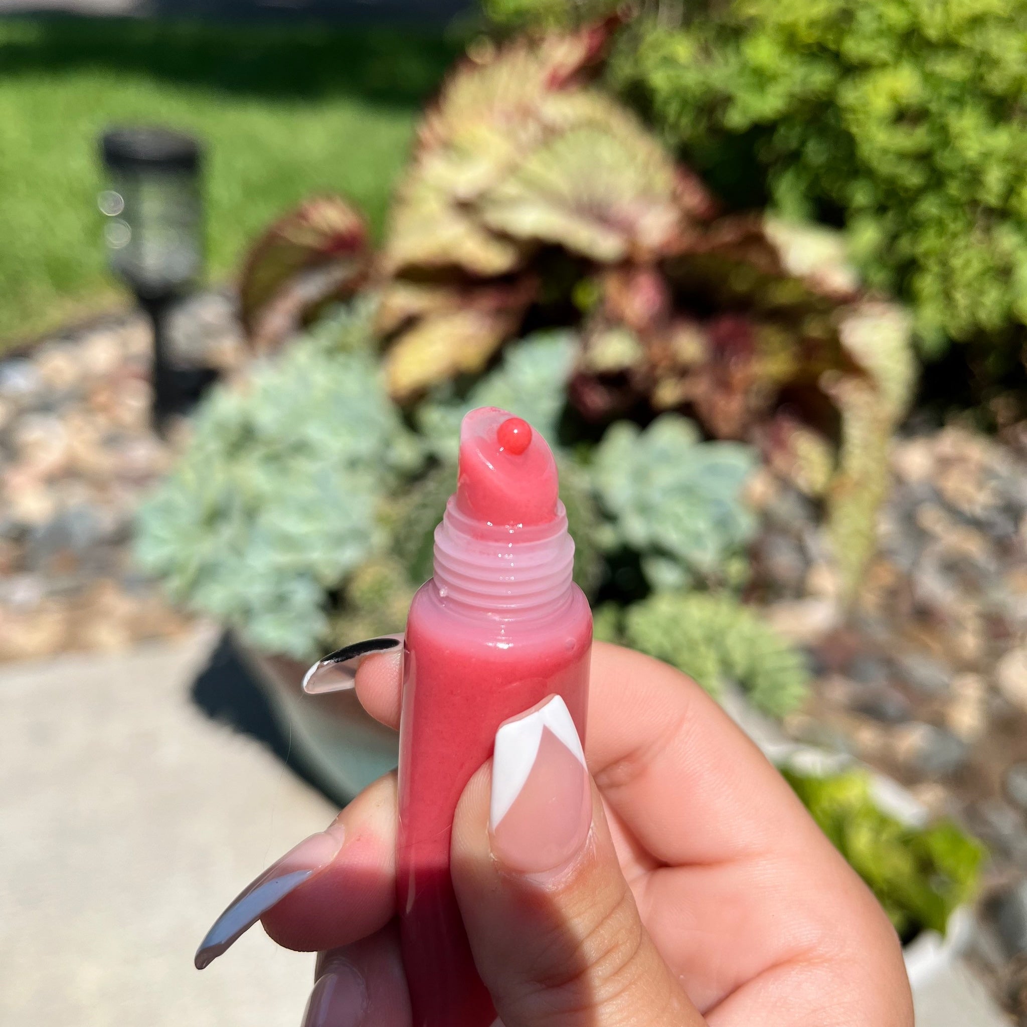 Lip gloss tube with pink liquid, held in hand with French manicure nails, outdoors in sunlight