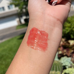 Lip gloss swatch, translucent red-pink color, shiny texture, applied on wrist