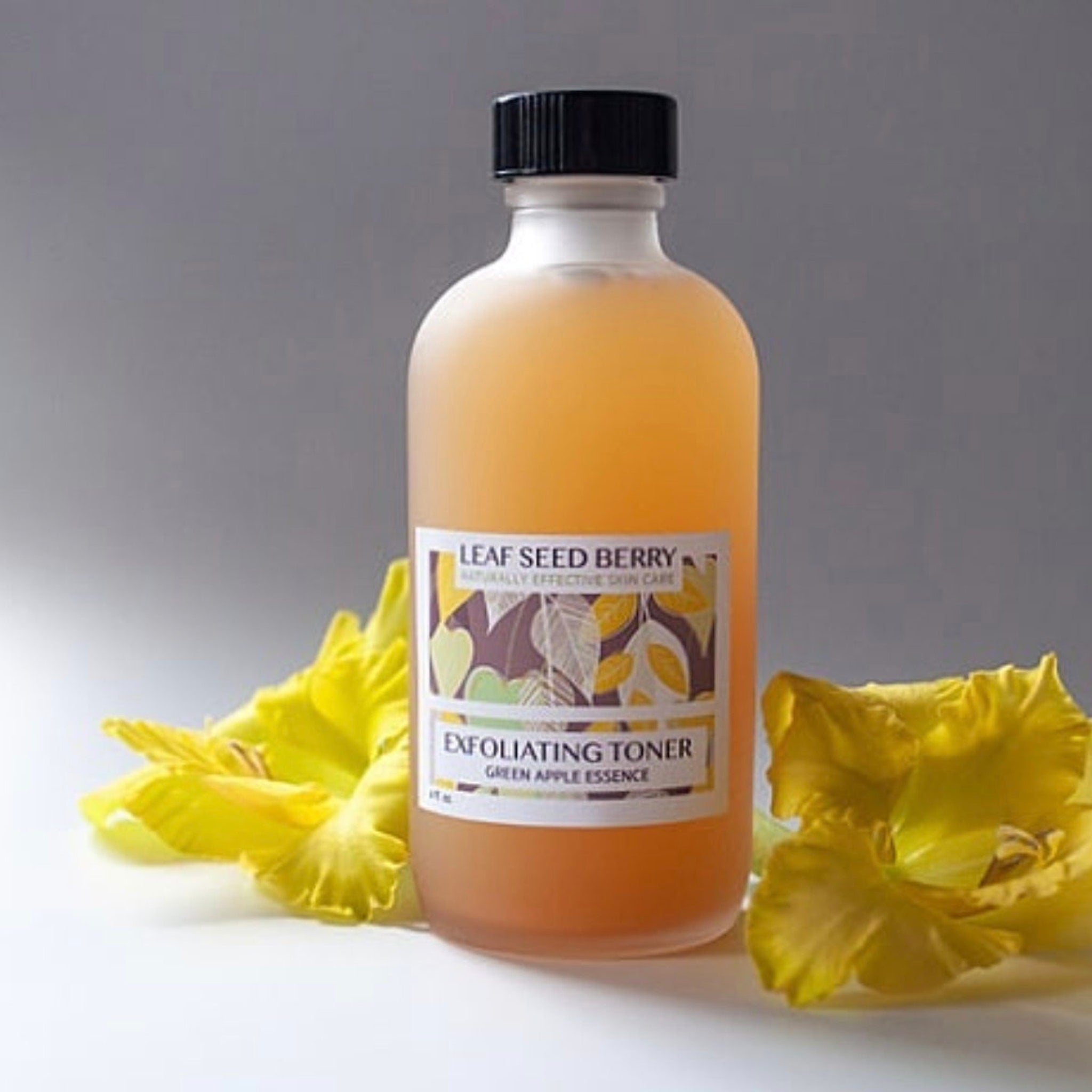 Exfoliating toner bottle with orange liquid, black cap, and yellow flower accents on white background