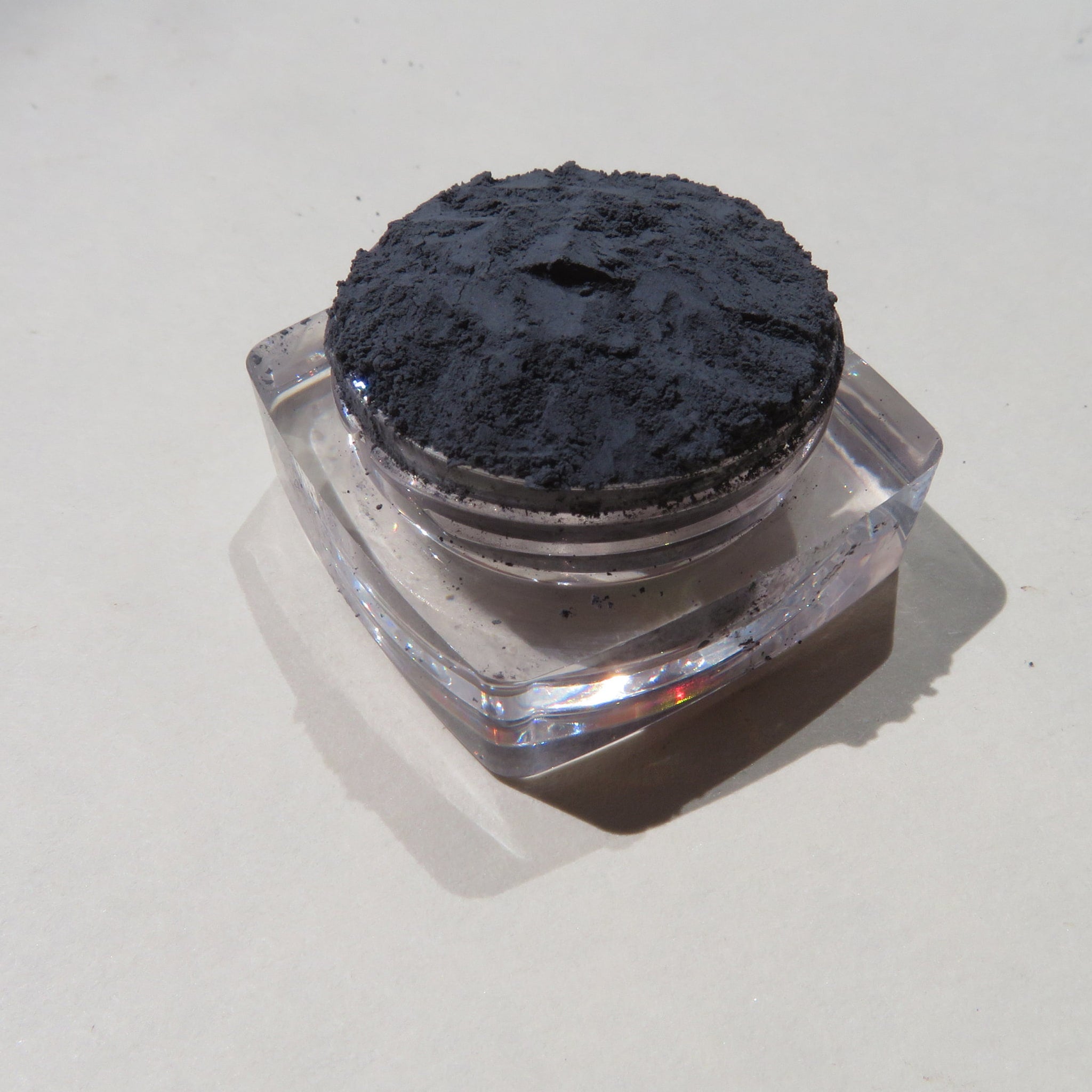 Powder pigment in dark gray, contained in clear square jar, on light surface