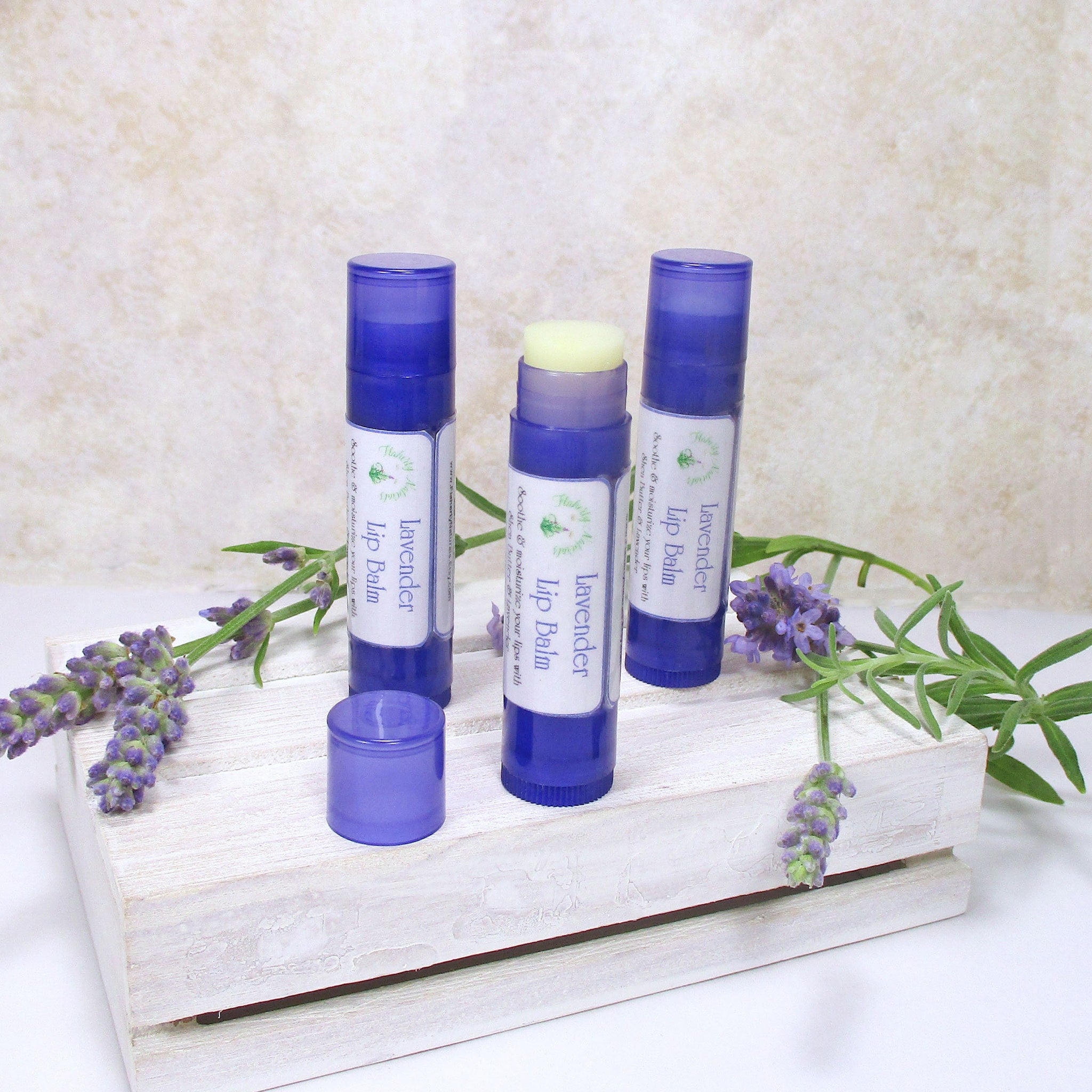 Lavender lip balm tubes in blue with white labels, on a white wooden box with lavender sprigs
