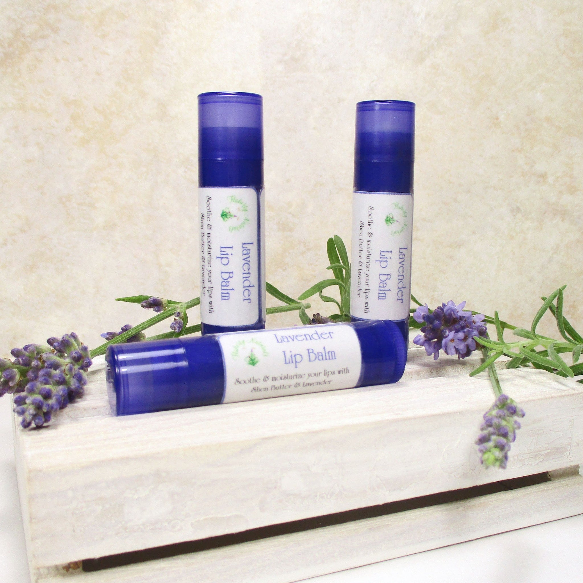 Lavender lip balm tubes, blue plastic, white label with text, and lavender sprigs on a white wooden box.