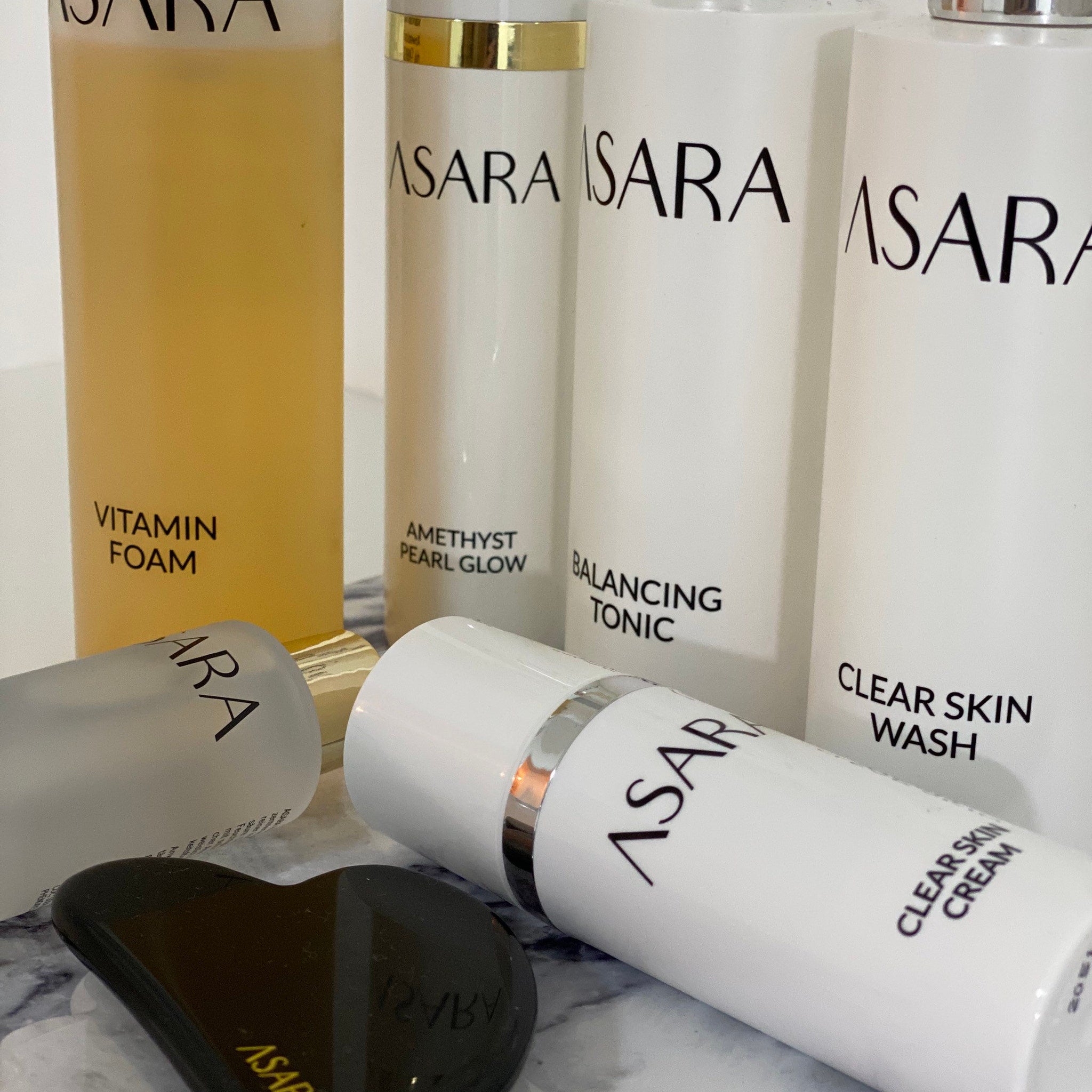 Skincare products in white bottles with black text, ASARA brand visible, and a black gua sha tool on a marble surface
