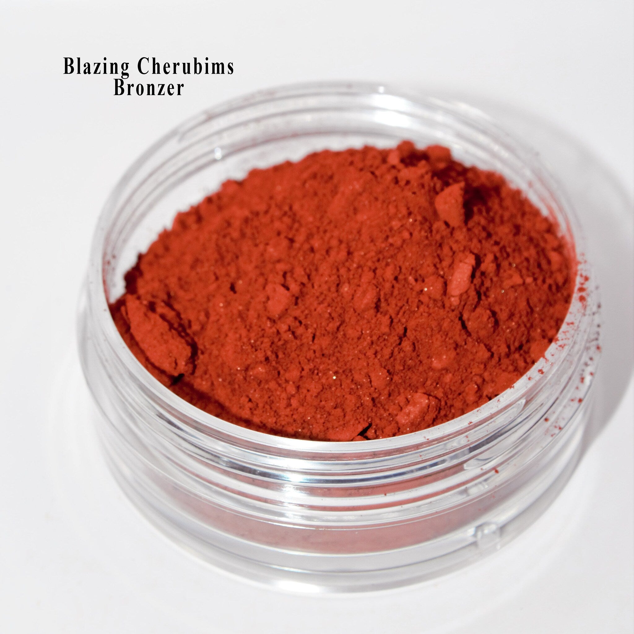 Bronzer powder in a clear plastic jar, with "Blazing Cherubims" text visible, red-orange color