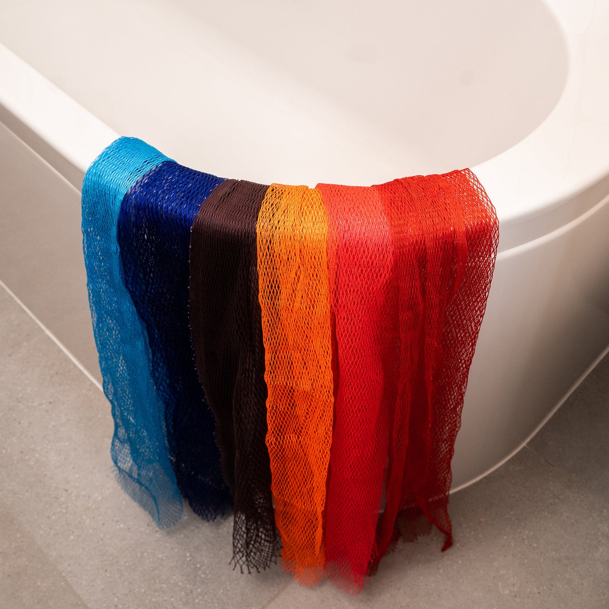 Bath loofahs in various colors, mesh material, hanging over a bathtub edge