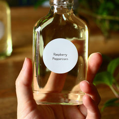 Clear glass bottle with yellow liquid, held by hand, white label says Raspberry Peppercorn
