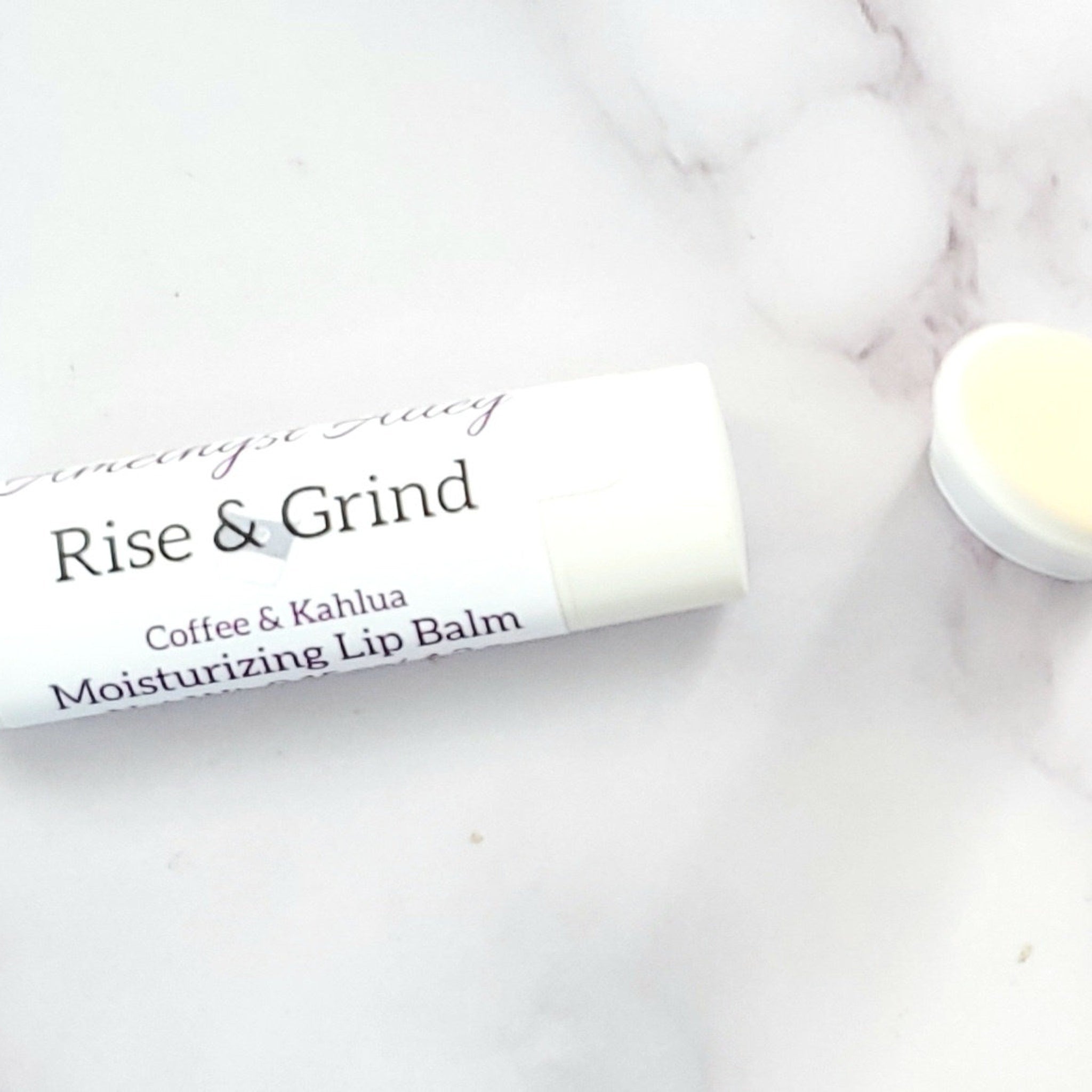 Lip balm tube, white with black text, "Rise & Grind" label, on a marble surface