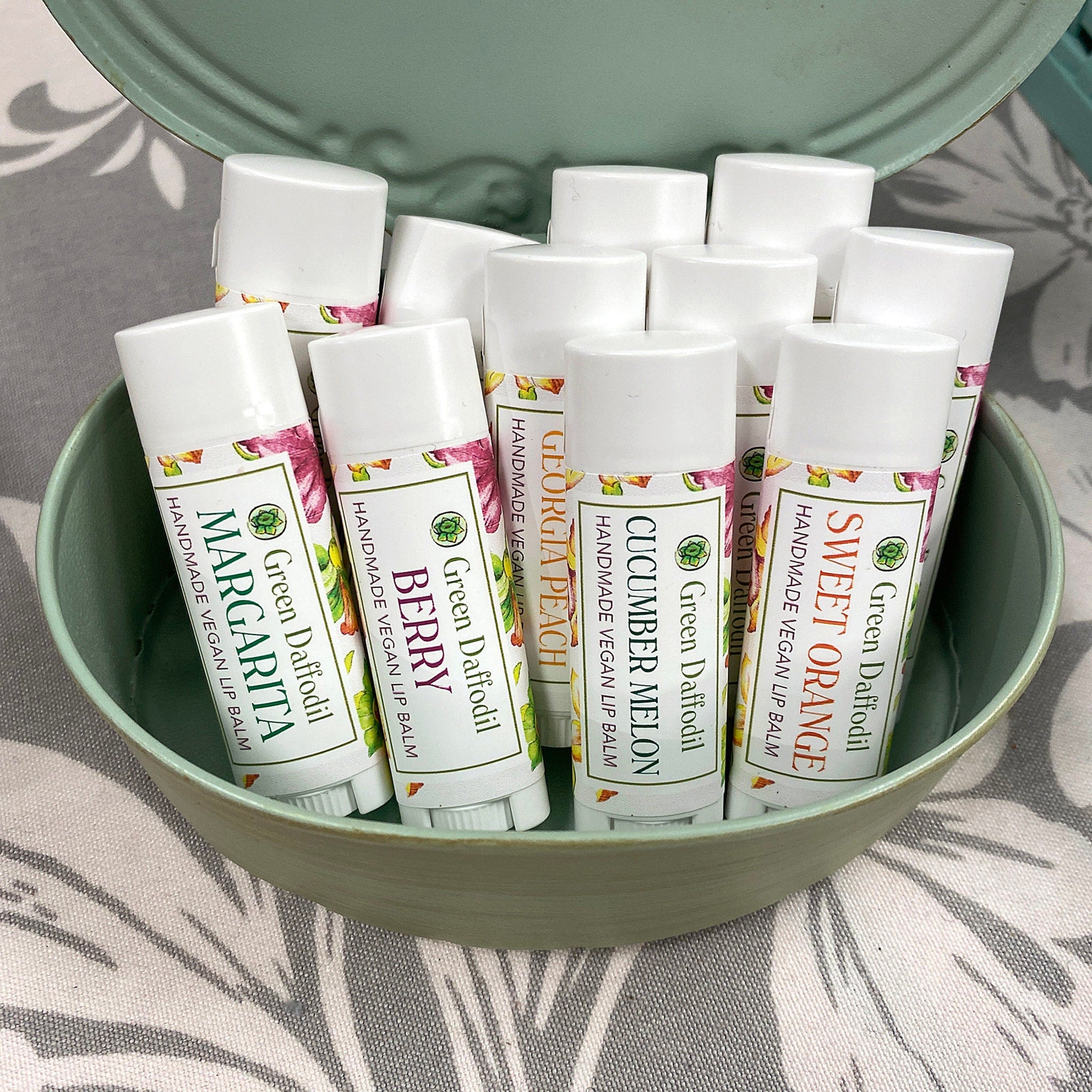 Lip balm tubes with white, printed labels in a green container on patterned fabric