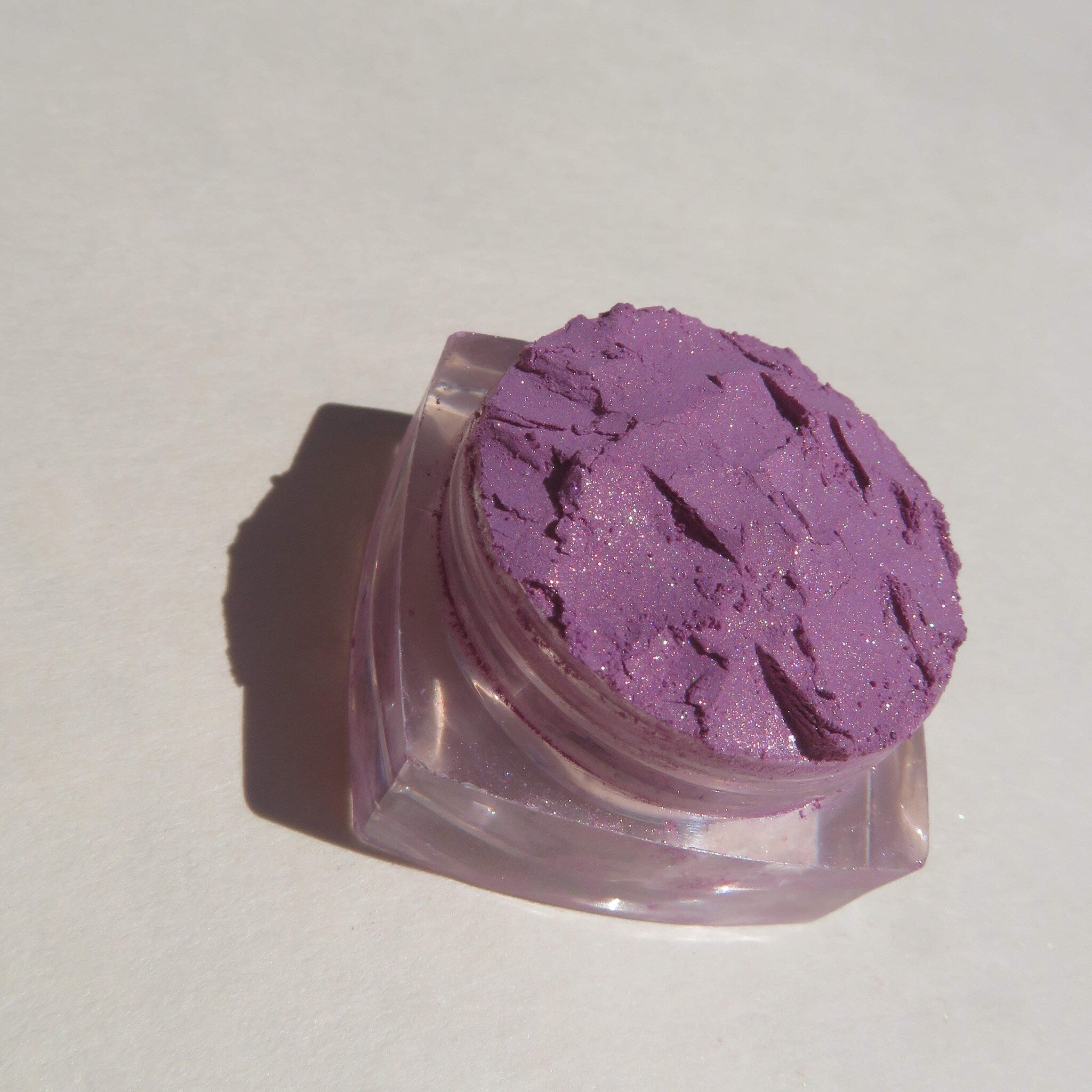 Side view of loose purple shimmer mineral eyeshadow in clear jar with silver lid, showcasing fine pigment texture
