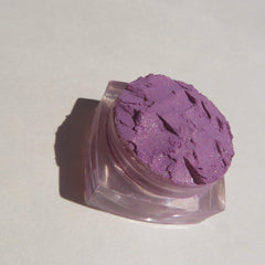 Side view of loose purple shimmer mineral eyeshadow in clear jar with silver lid, showcasing fine pigment texture