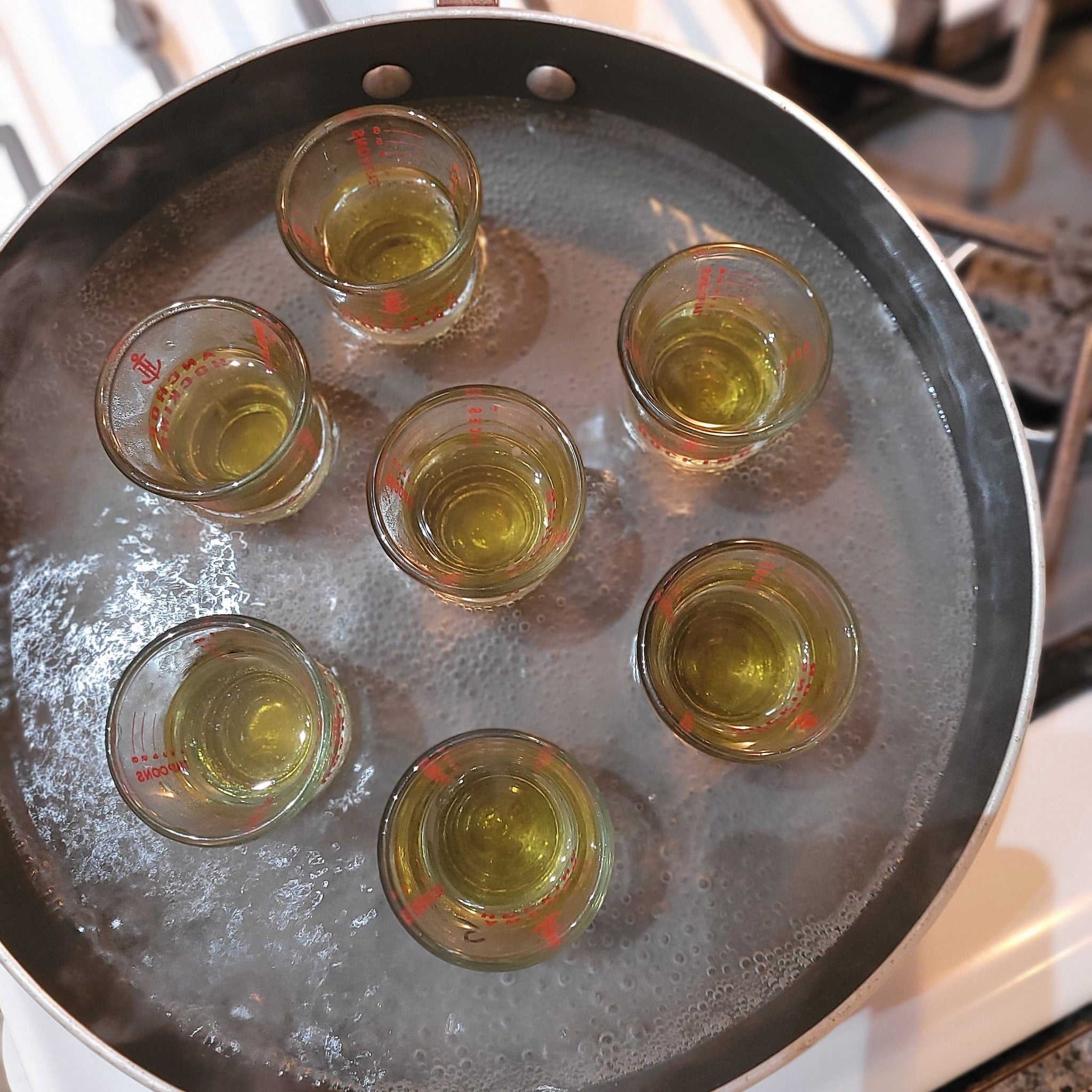 Small glass measuring cups with yellow liquid in a pot of boiling water