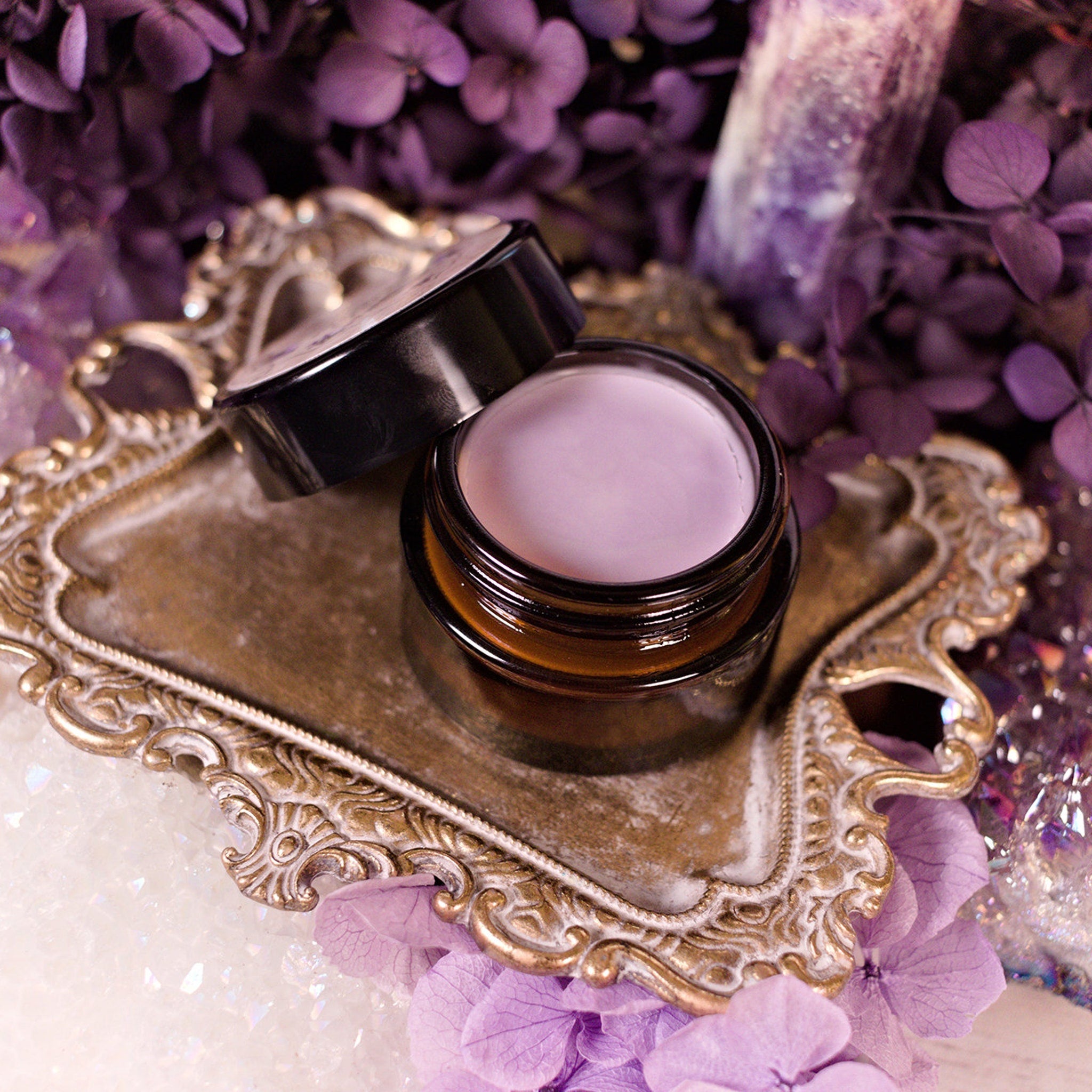 Cosmetic jar with light purple cream, brown glass, and black lid on ornate gold tray