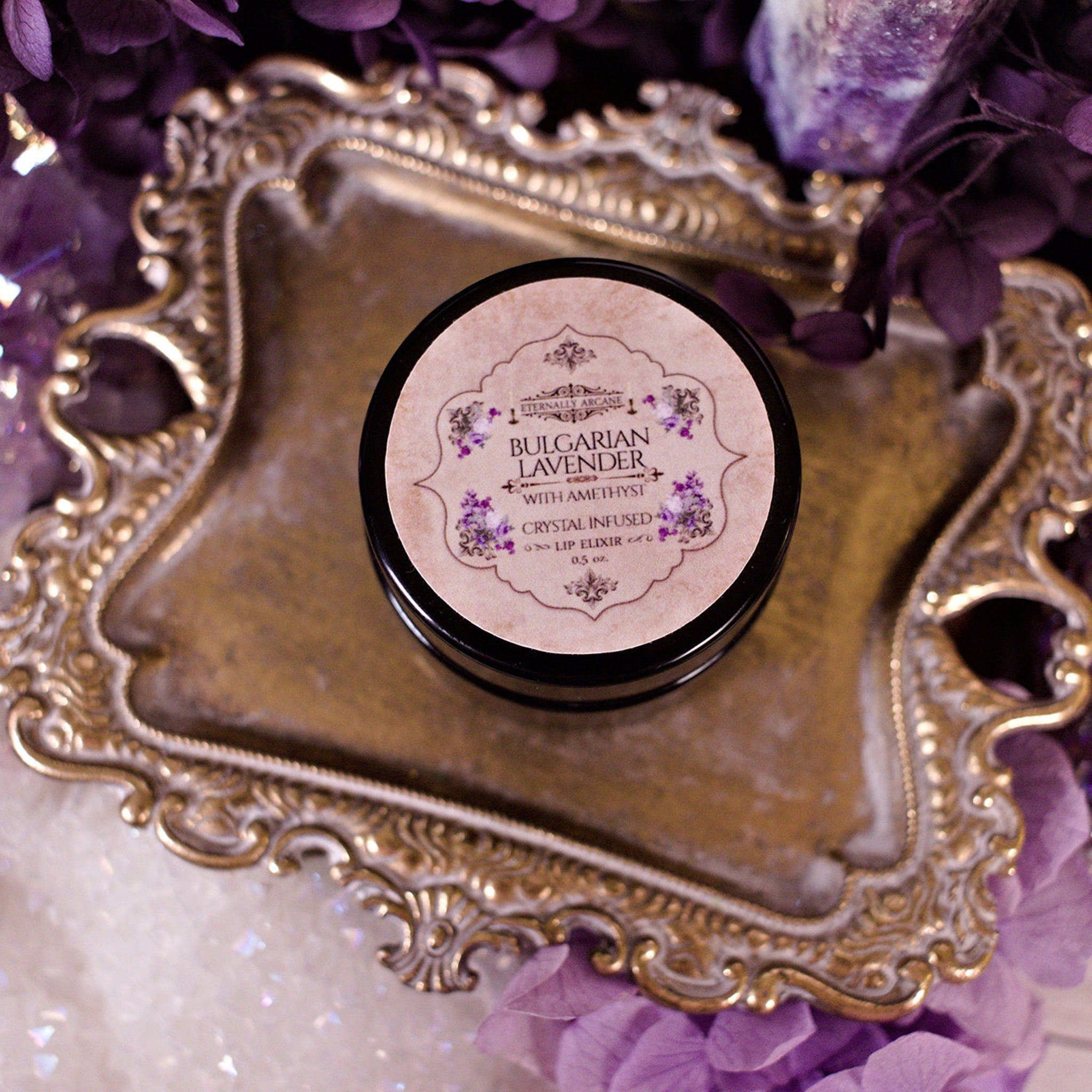 Lip elixir in black tin with beige label, gold ornate tray, and purple flowers