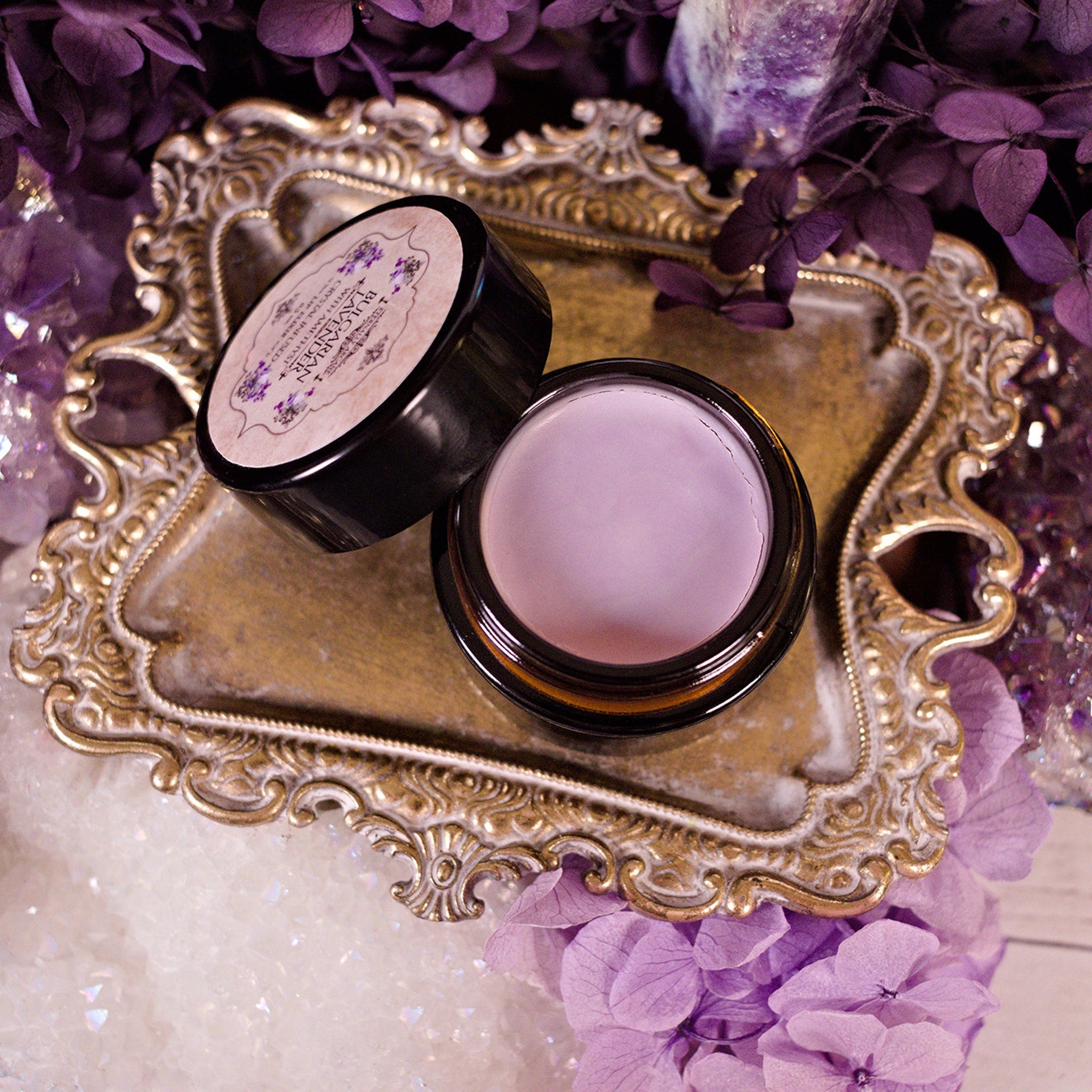 Cosmetic jar with black rim and light purple cream on ornate gold tray with flowers