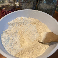 Powdered substance in white bowl, beige color, wooden spoon, with jars of herbs in background