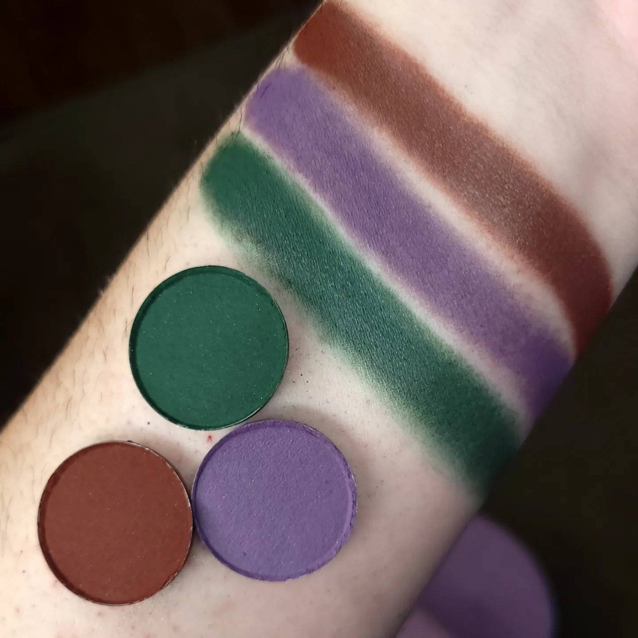 Eyeshadow swatches in green, purple, and brown shades on arm, circular pans shown