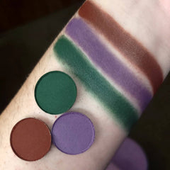 Eyeshadow swatches in green, purple, and brown shades on arm, circular pans shown