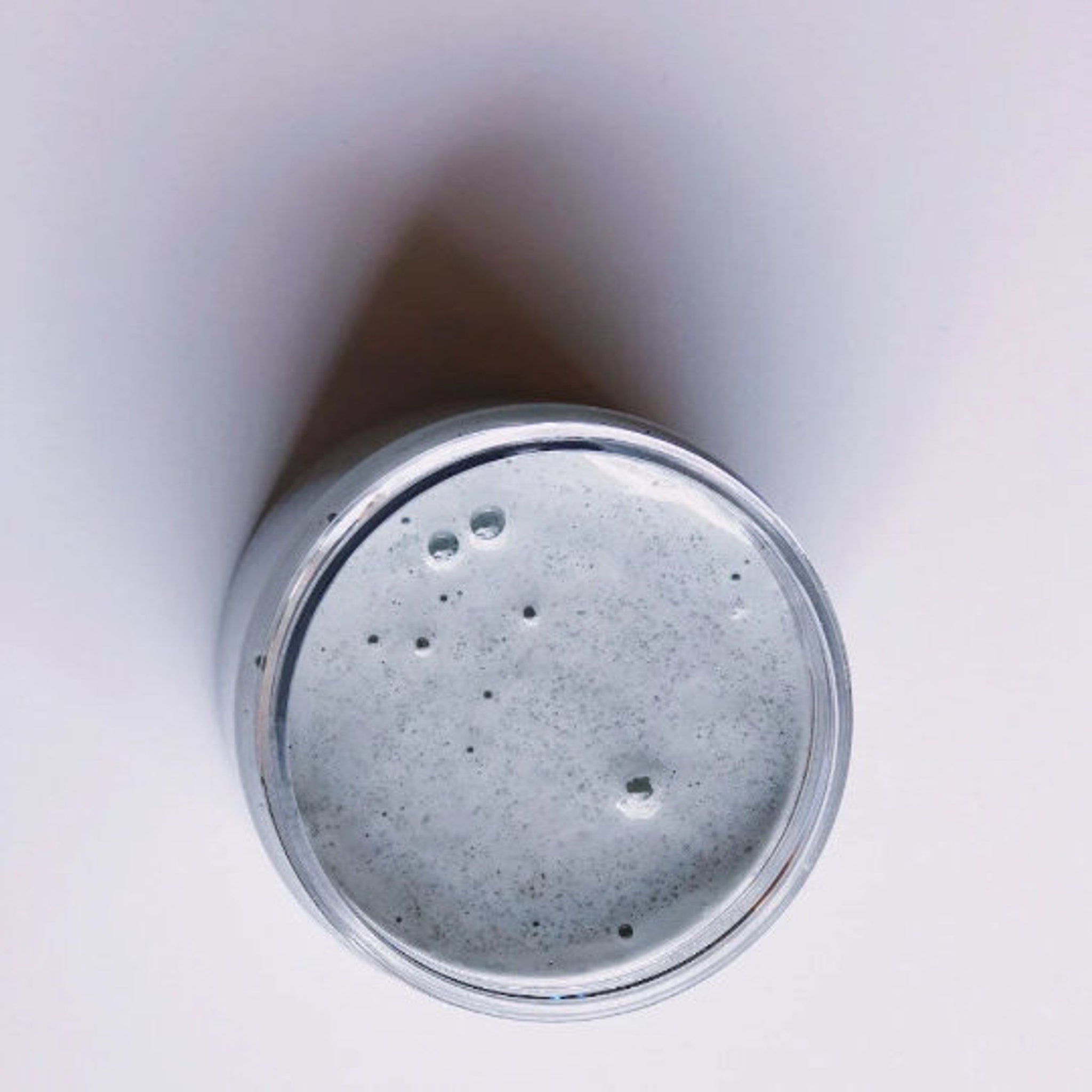 Jar of greyish-blue substance with clear container, small dark specks, and smooth texture