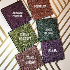 Side angle of pressed trichrome eyeshadow pan showing color-shifting shimmer from violet to emerald green