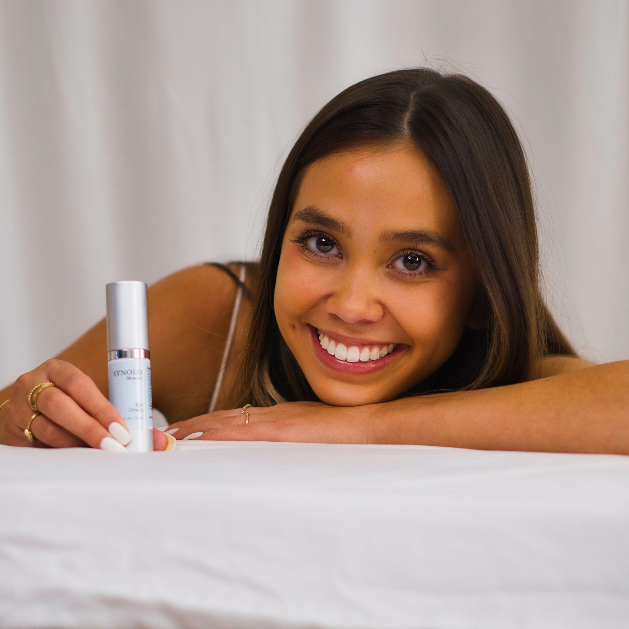 Eye cream bottle, silver and white, held by woman with white nails, SYNOLO skincare brand visible